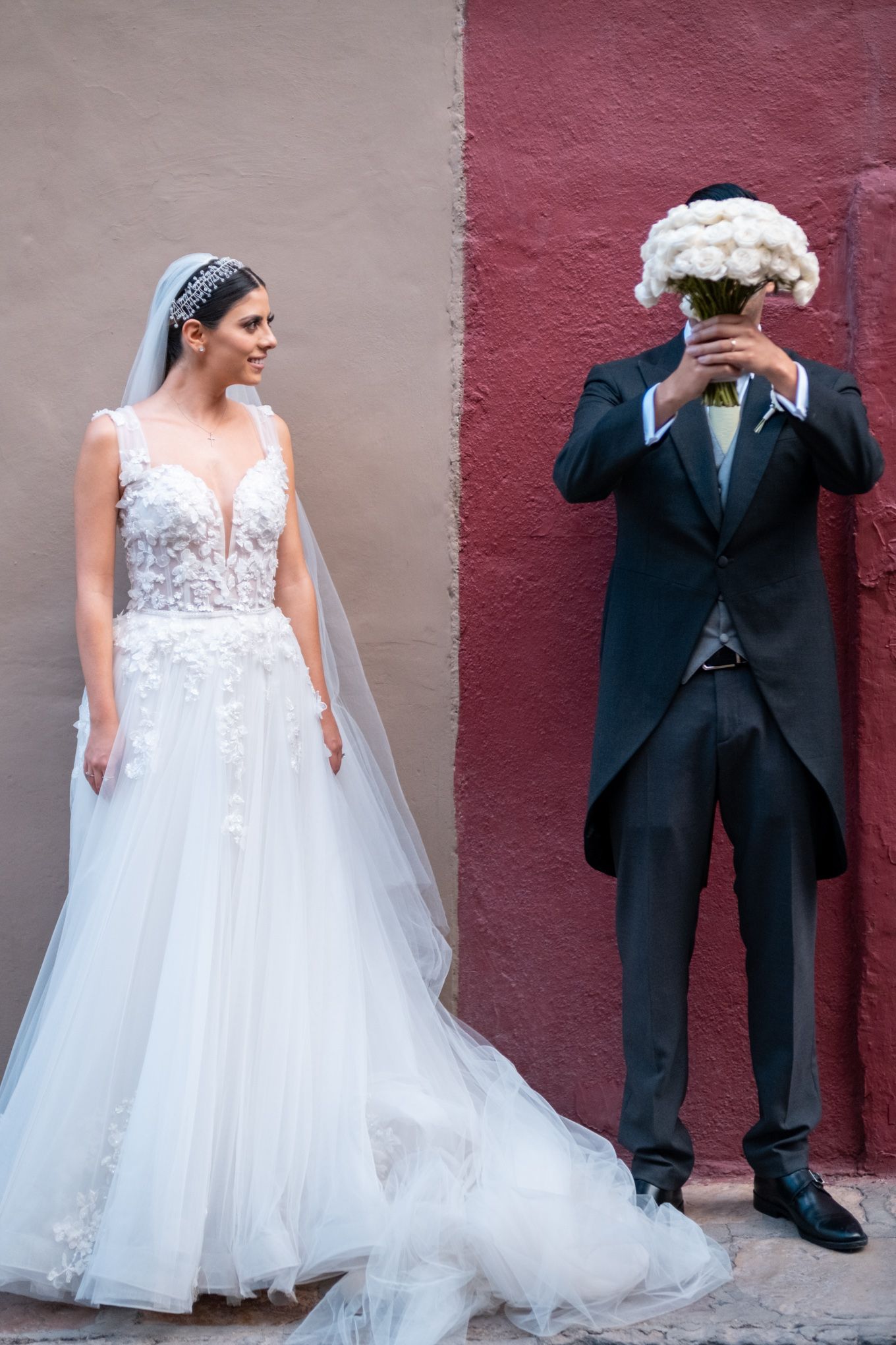 Playful wedding portrait against a two-toned wall with a bouquet hiding the groom's face.