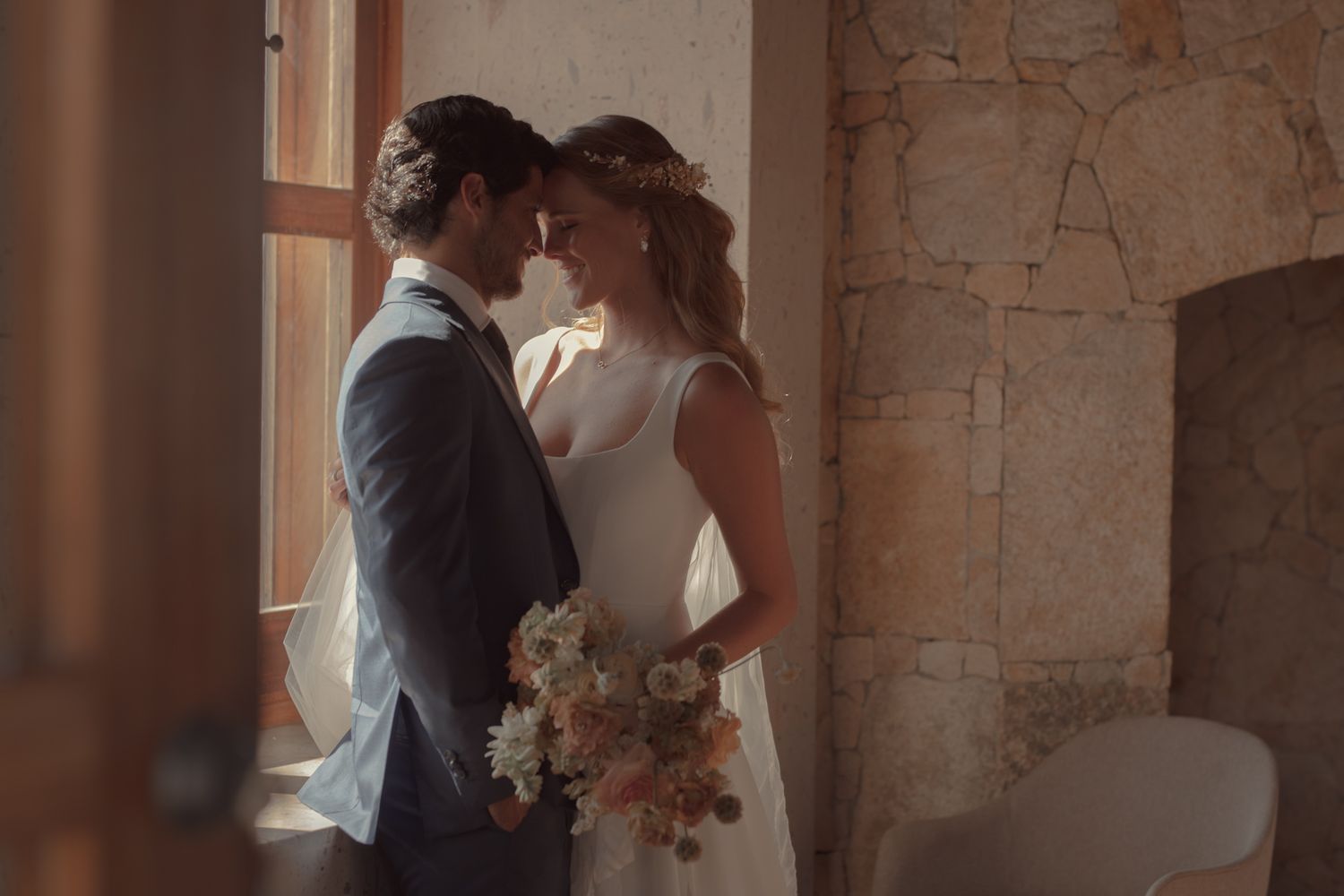 A romantic moment between newlyweds standing by a sunlit window with a bridal bouquet.