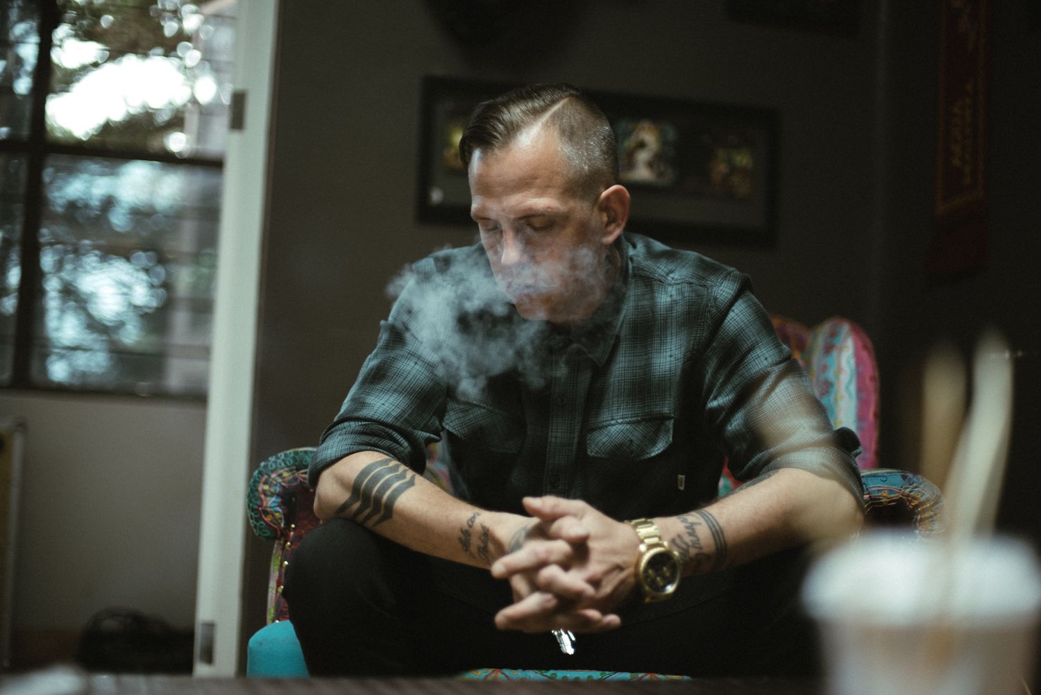 A person in a plaid shirt sits pensively in a dimly lit room with smoke lingering in the air.