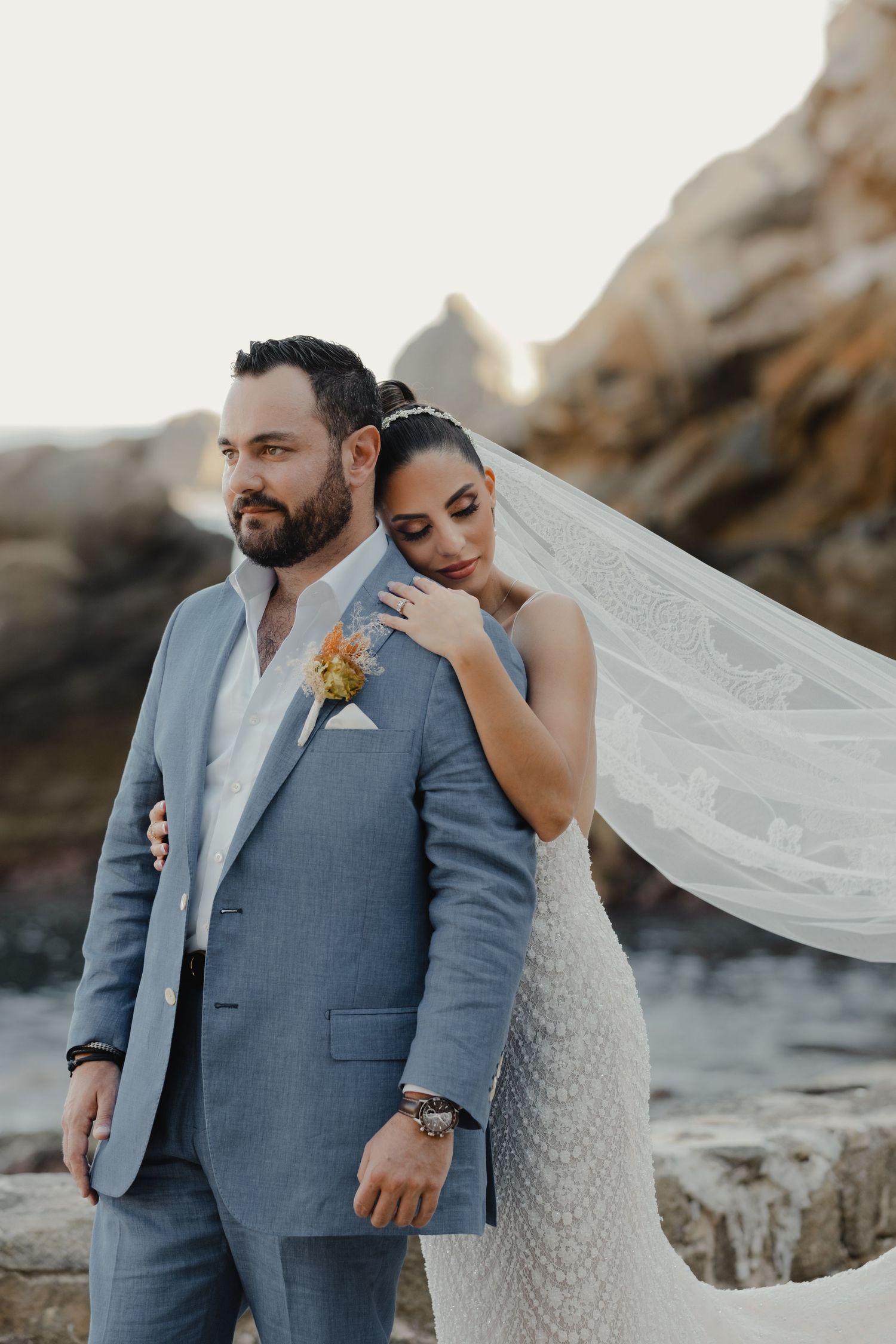 Bride and groom embrace on rocky coastline at sunset, with flowing veil and grey suit against natural backdrop.