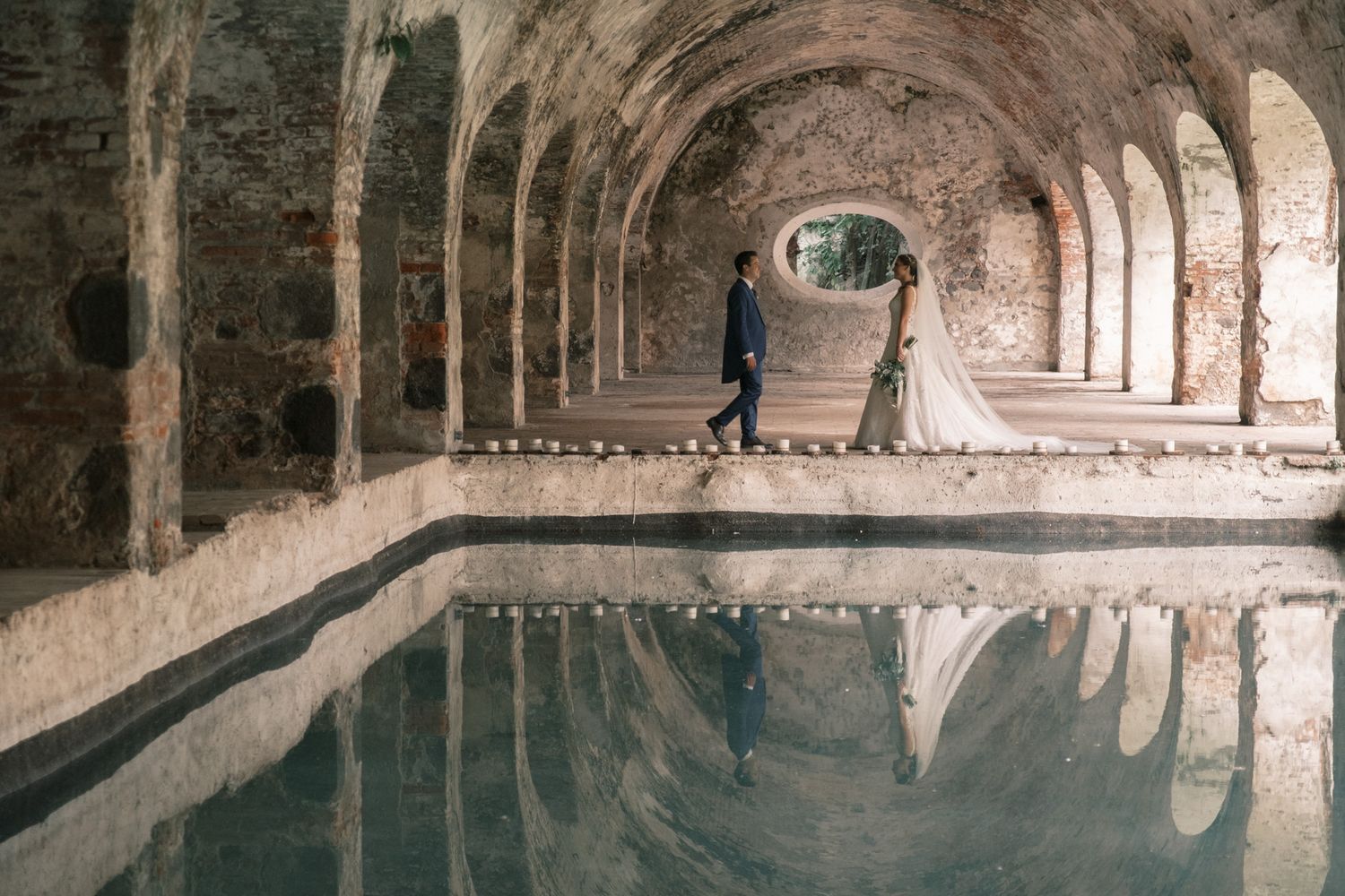 Ancient stone arched corridor with reflective indoor pool showing architectural details and symmetrical reflections.
