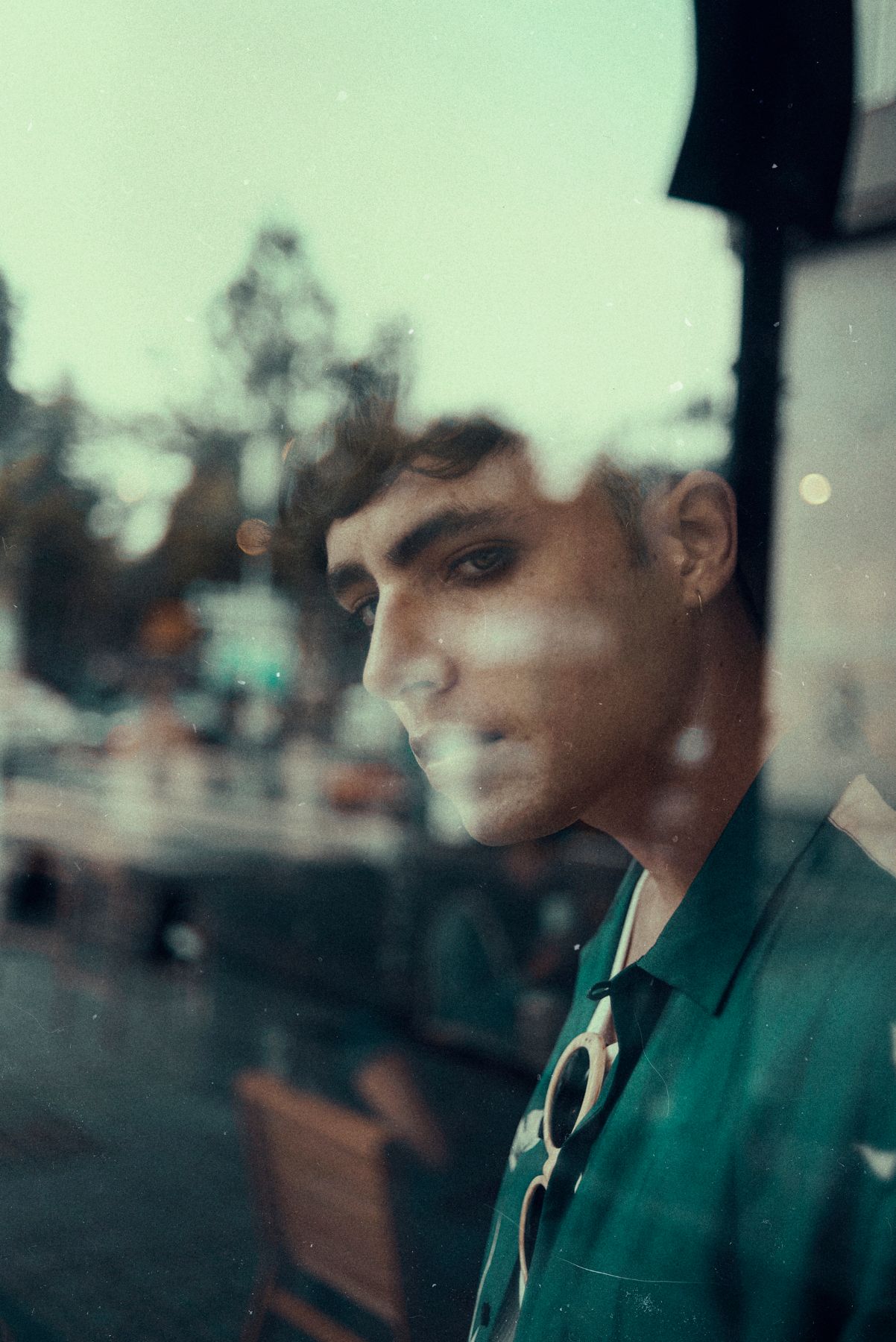 Moody portrait through window reflection with soft urban bokeh in background.