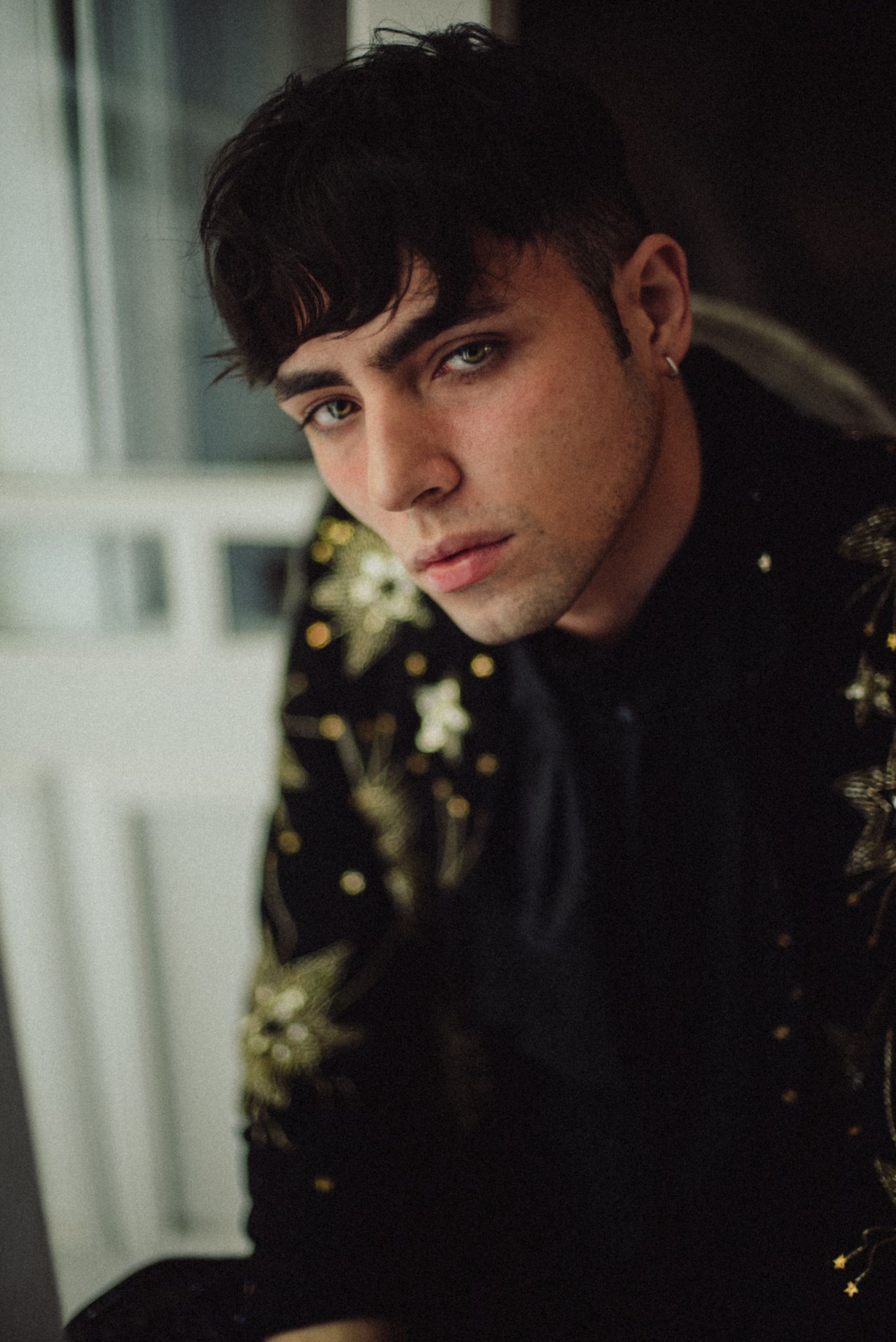 Moody portrait of person in black turtleneck against window with dramatic lighting and foliage background.