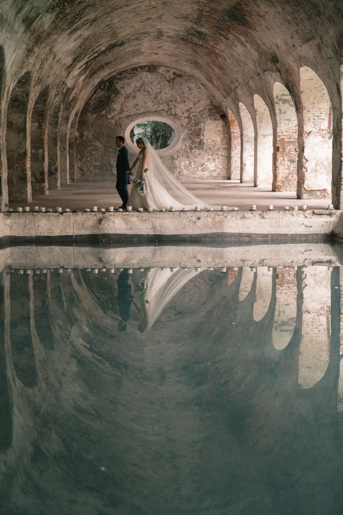 A wedding couple stands in an ancient stone archway reflected in still water below creating a mirror image.