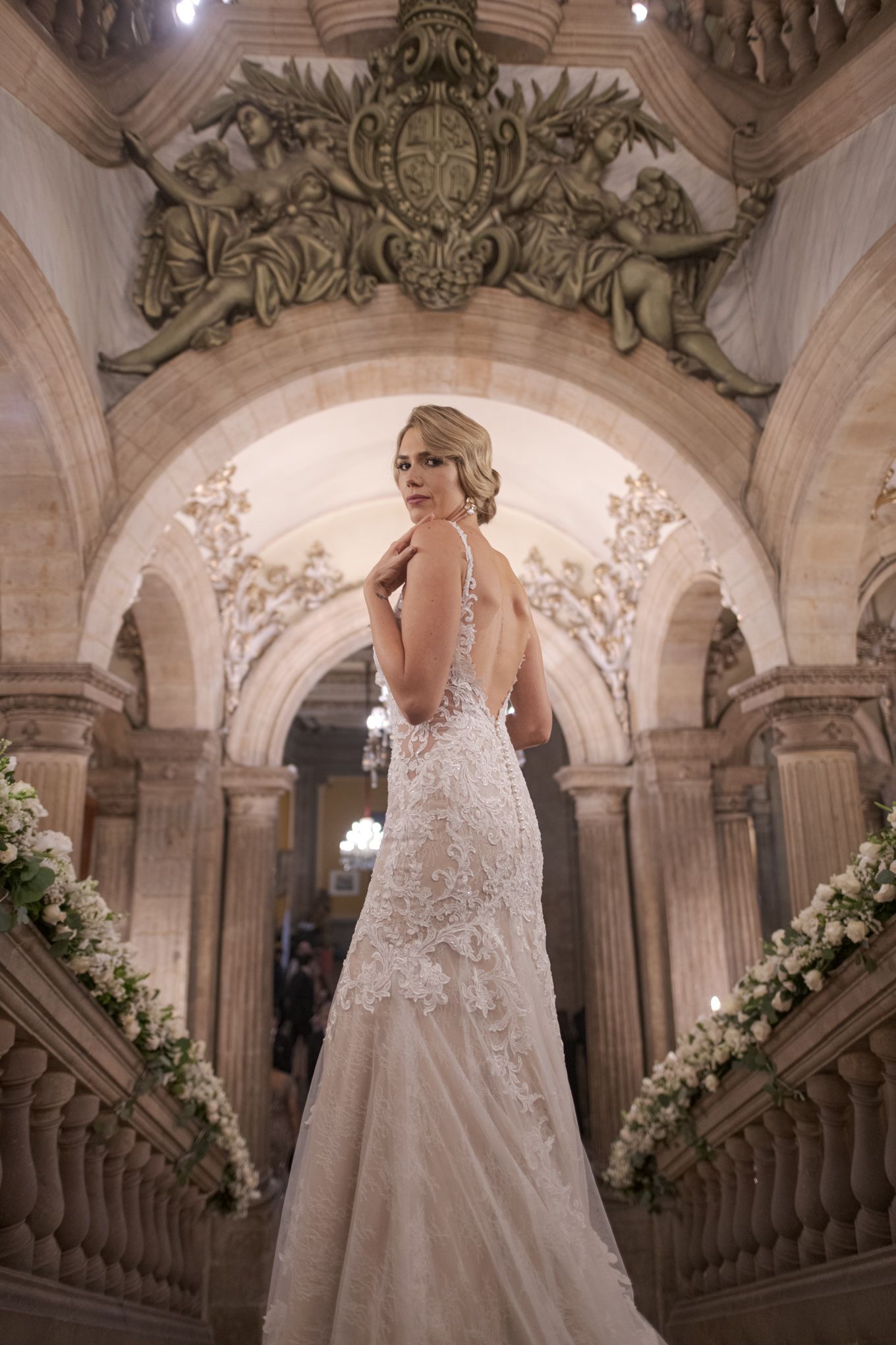 Bride in lace gown poses in ornate stone archway decorated with white floral arrangements.