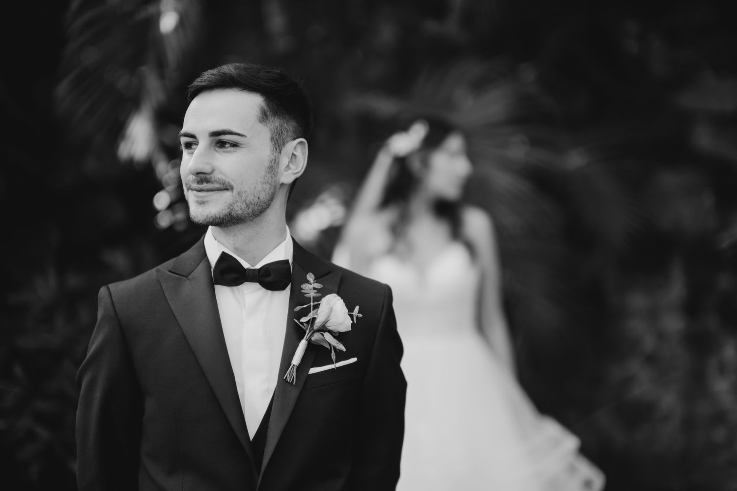 Black and white portrait of a groom in tuxedo with boutonniere against blurred background.