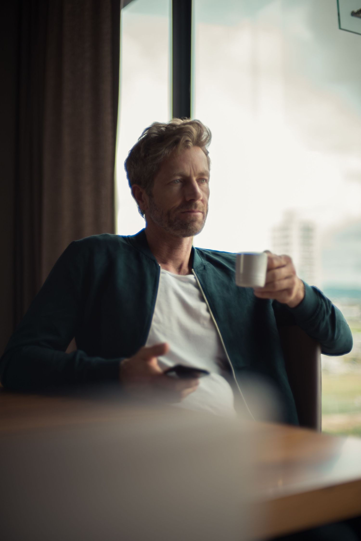 Casual portrait of person enjoying coffee by window with cityscape view.