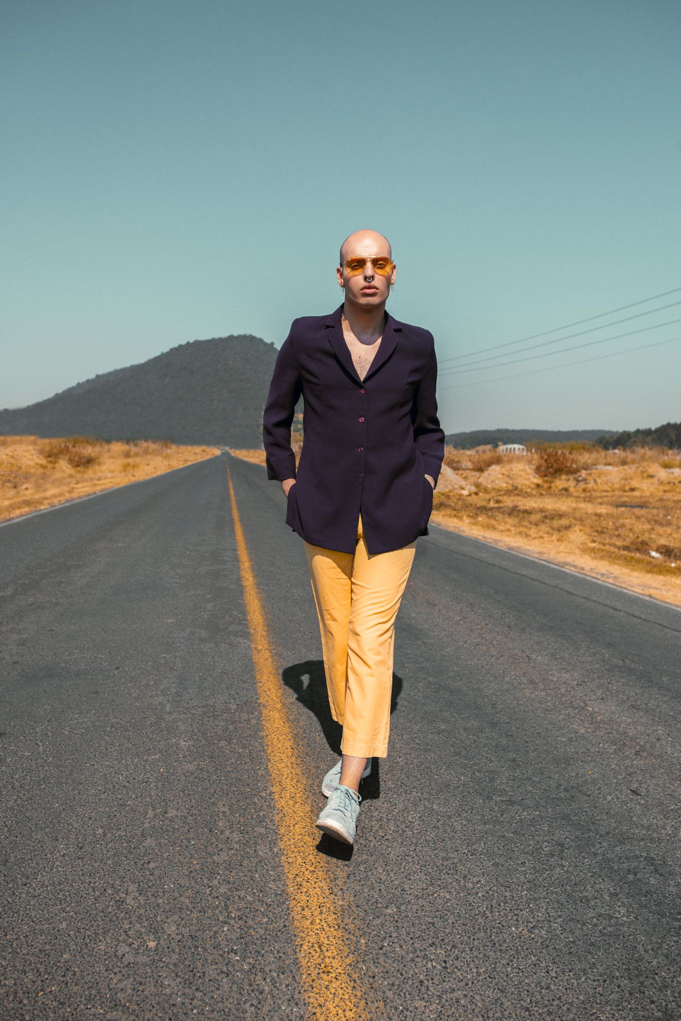 Stylish figure in purple blazer and yellow pants walking on an empty desert road with mountains in background.