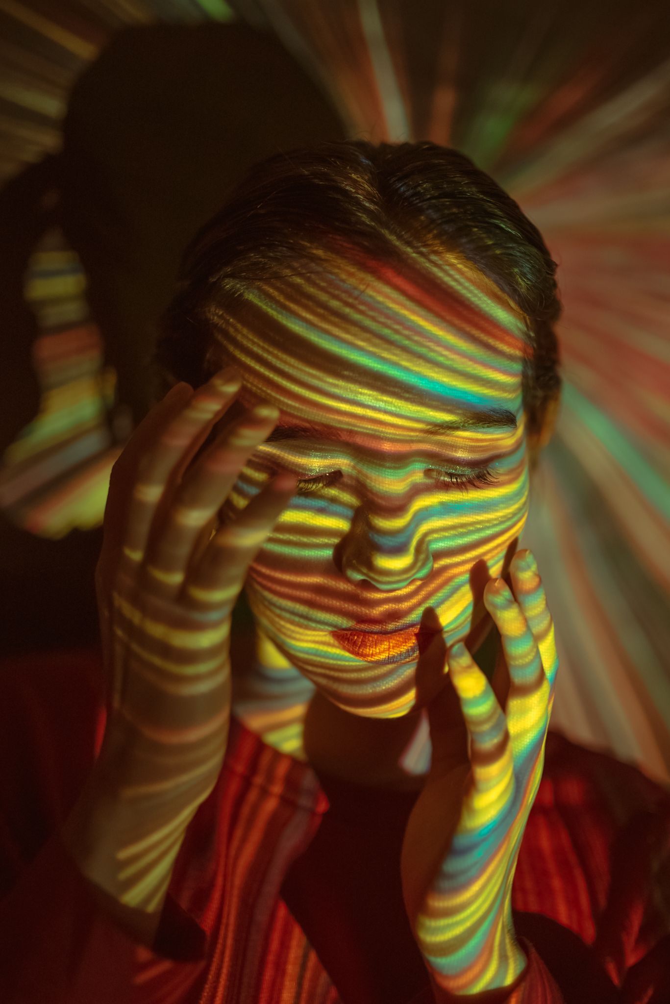 Artistic photograph showing rainbow light patterns projected onto a figure covering their face with hands.