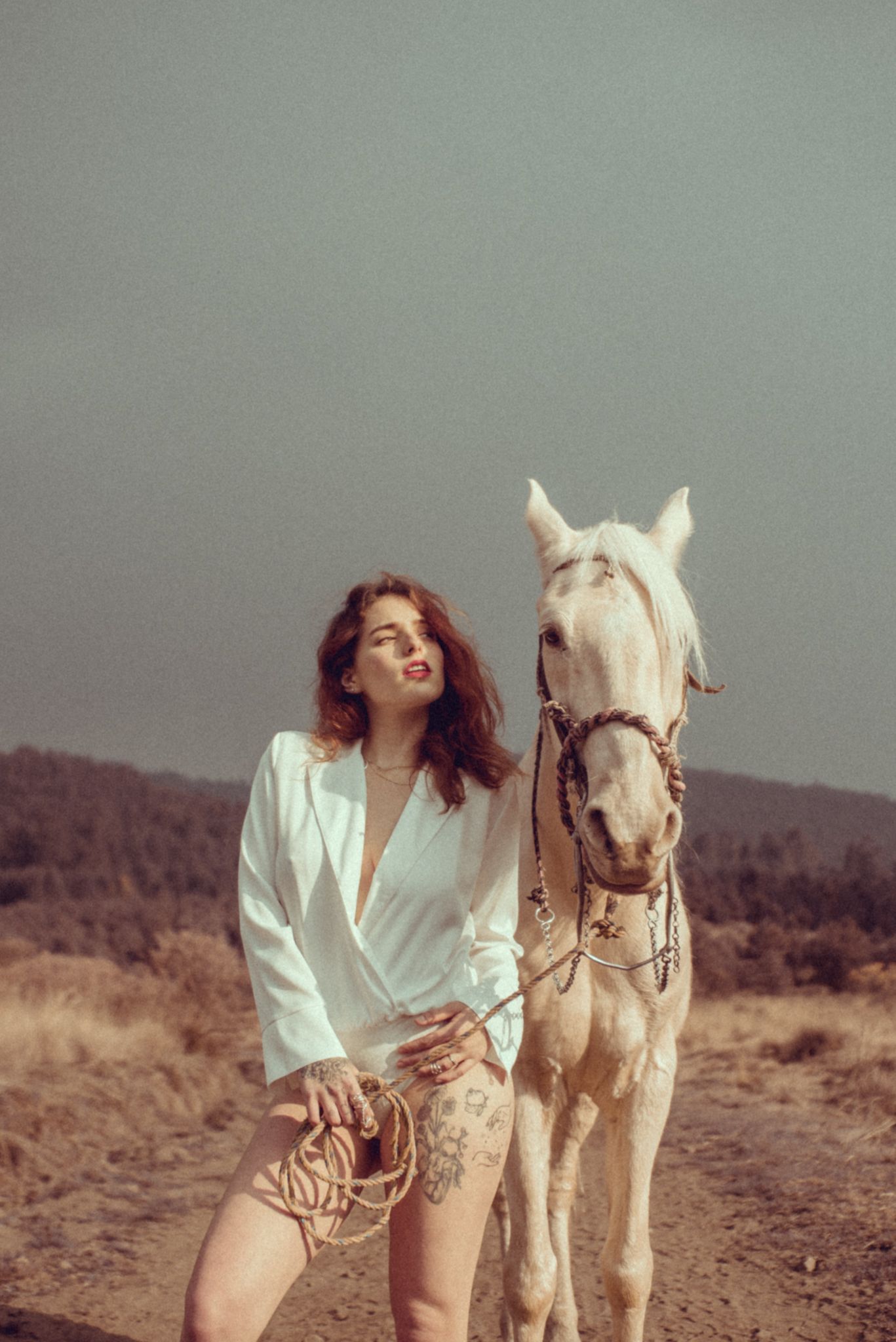 A person smiles while standing next to a white horse in rustic outdoor scenery.