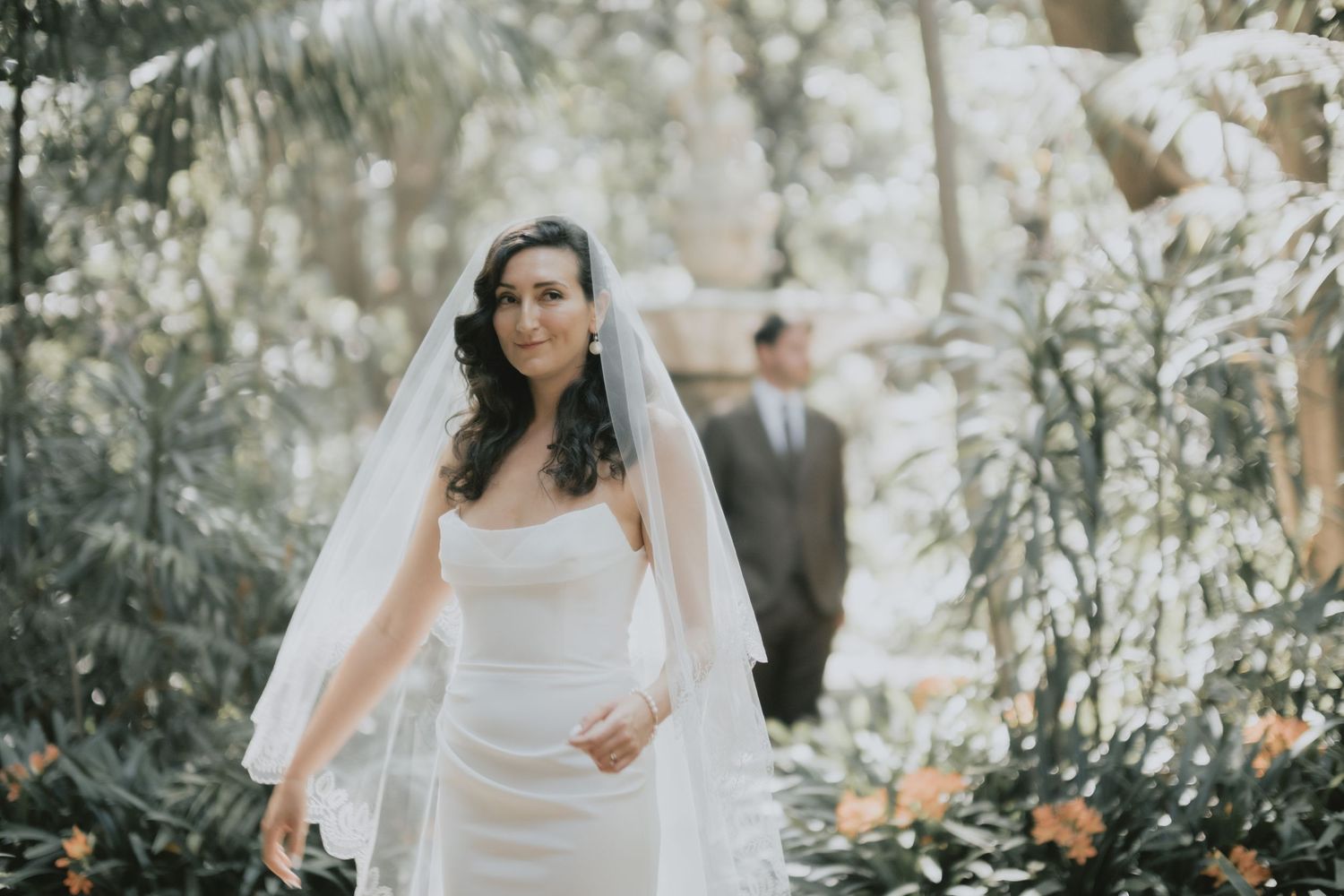A bride in a white gown walks through a lush garden setting with long flowing veil.
