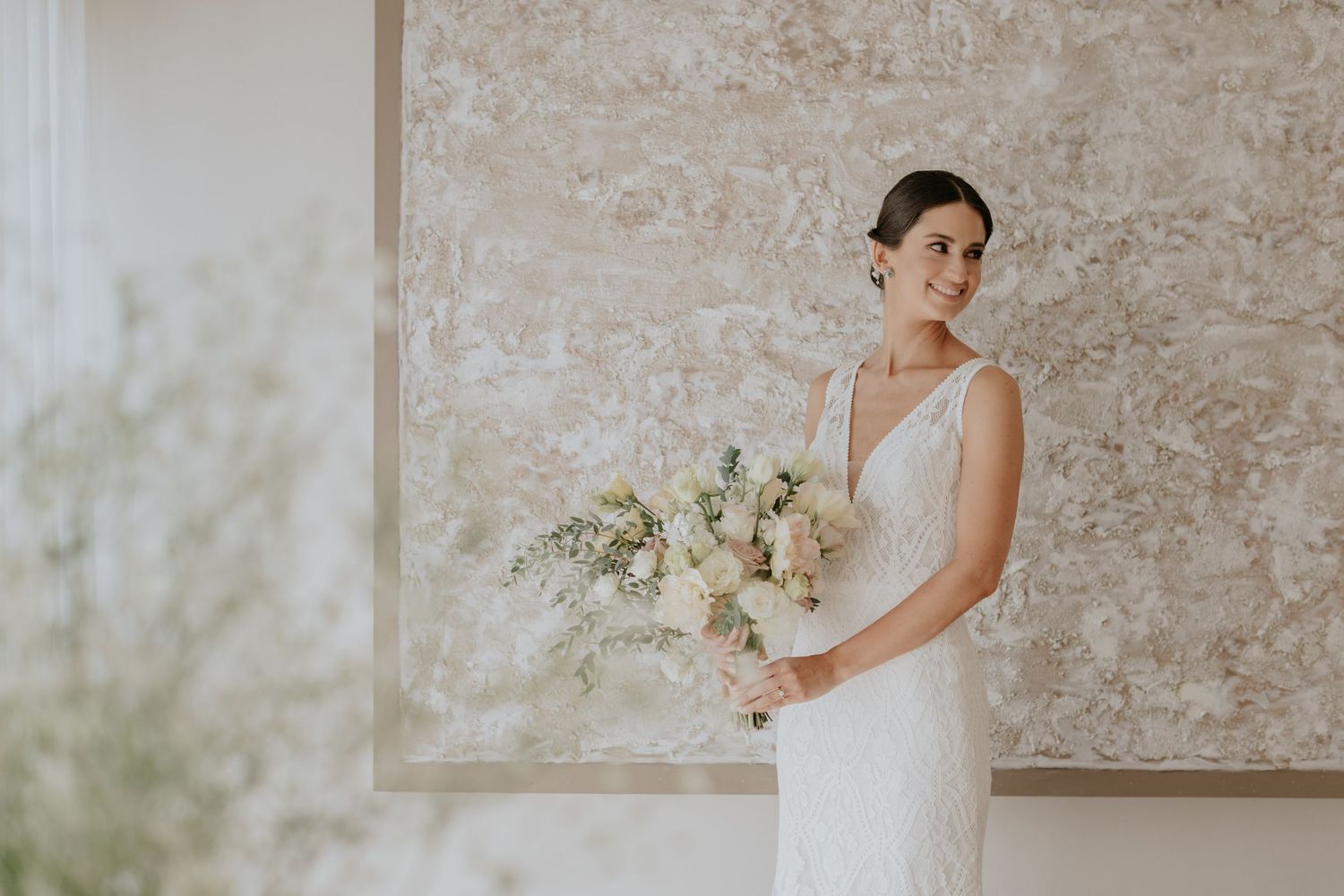 Bride in v-neck wedding dress holds white and green bouquet against textured wall.
