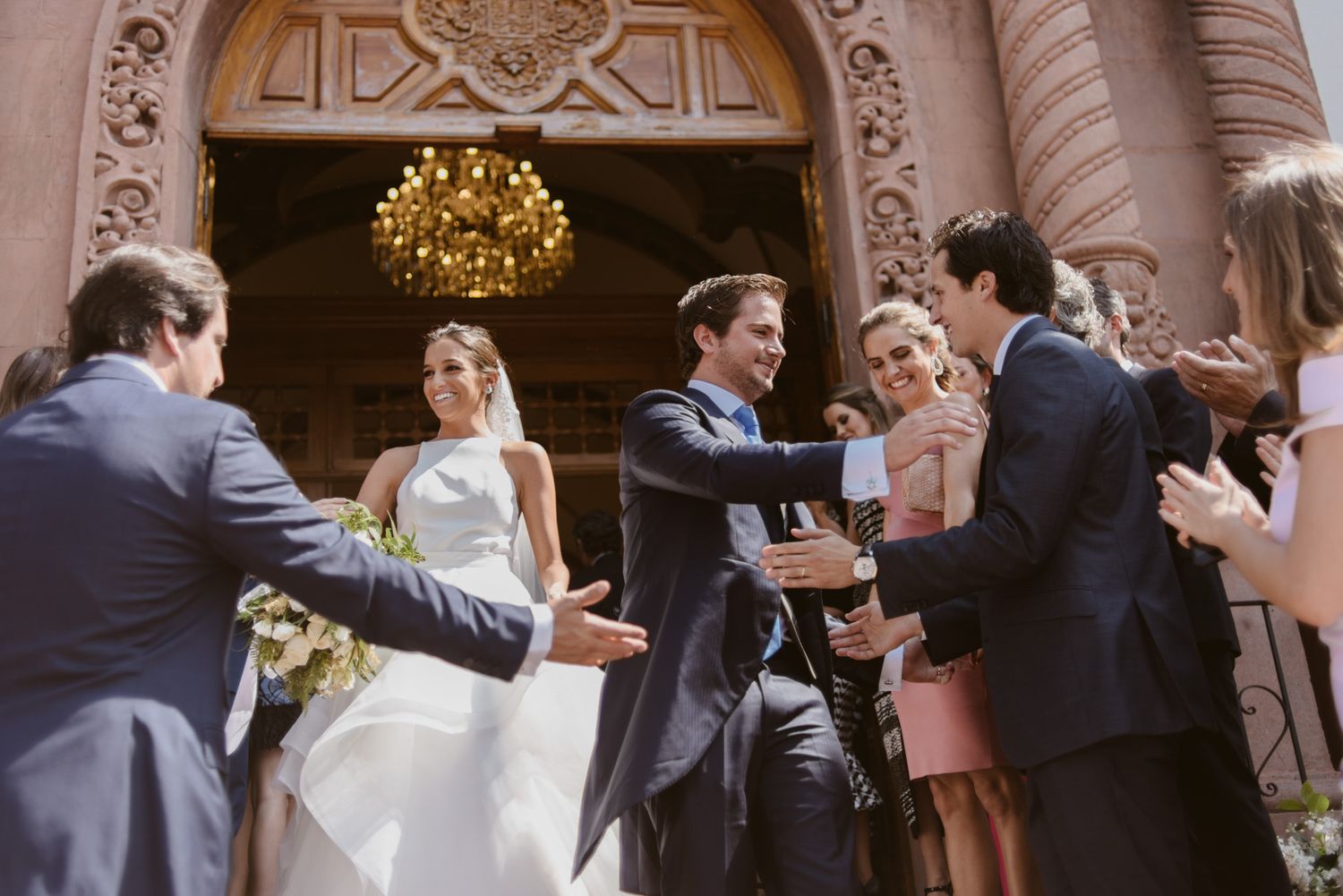 Wedding party gathers on ornate steps outside venue with classic architecture and chandelier.