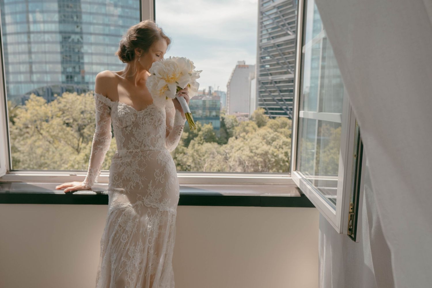A bride in a fitted lace gown looks out over a cityscape from a modern high-rise window.