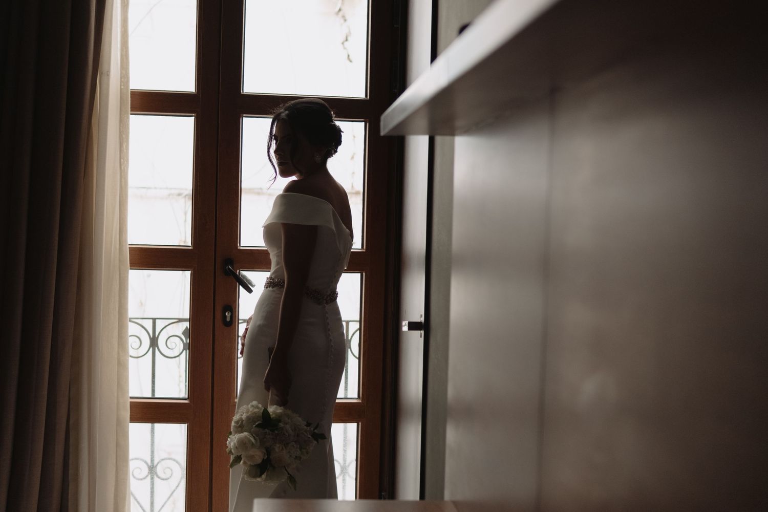 Silhouette of person in wedding dress looking through window in morning light.