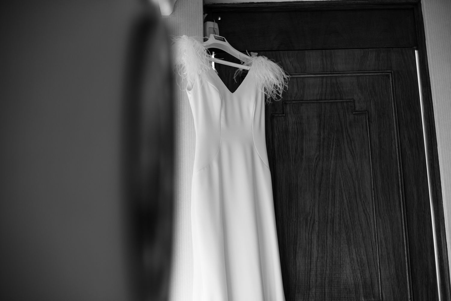 Minimalist black and white photo of white wedding dress hanging on dark wood door.