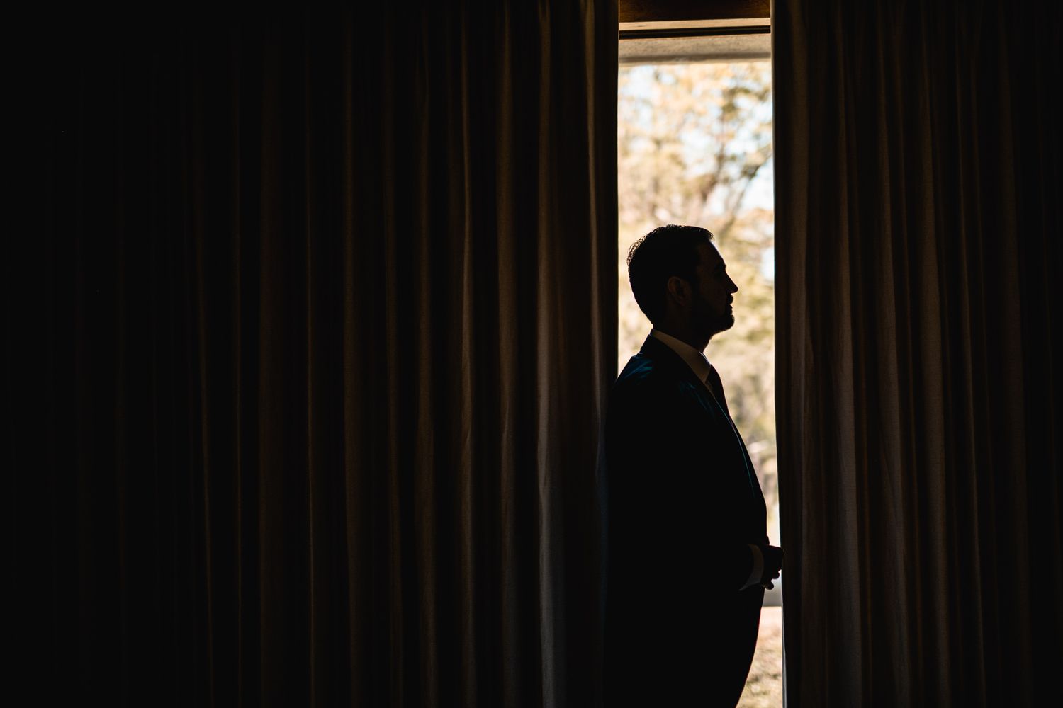 A silhouetted figure stands between dark curtains gazing at bright light from a window.