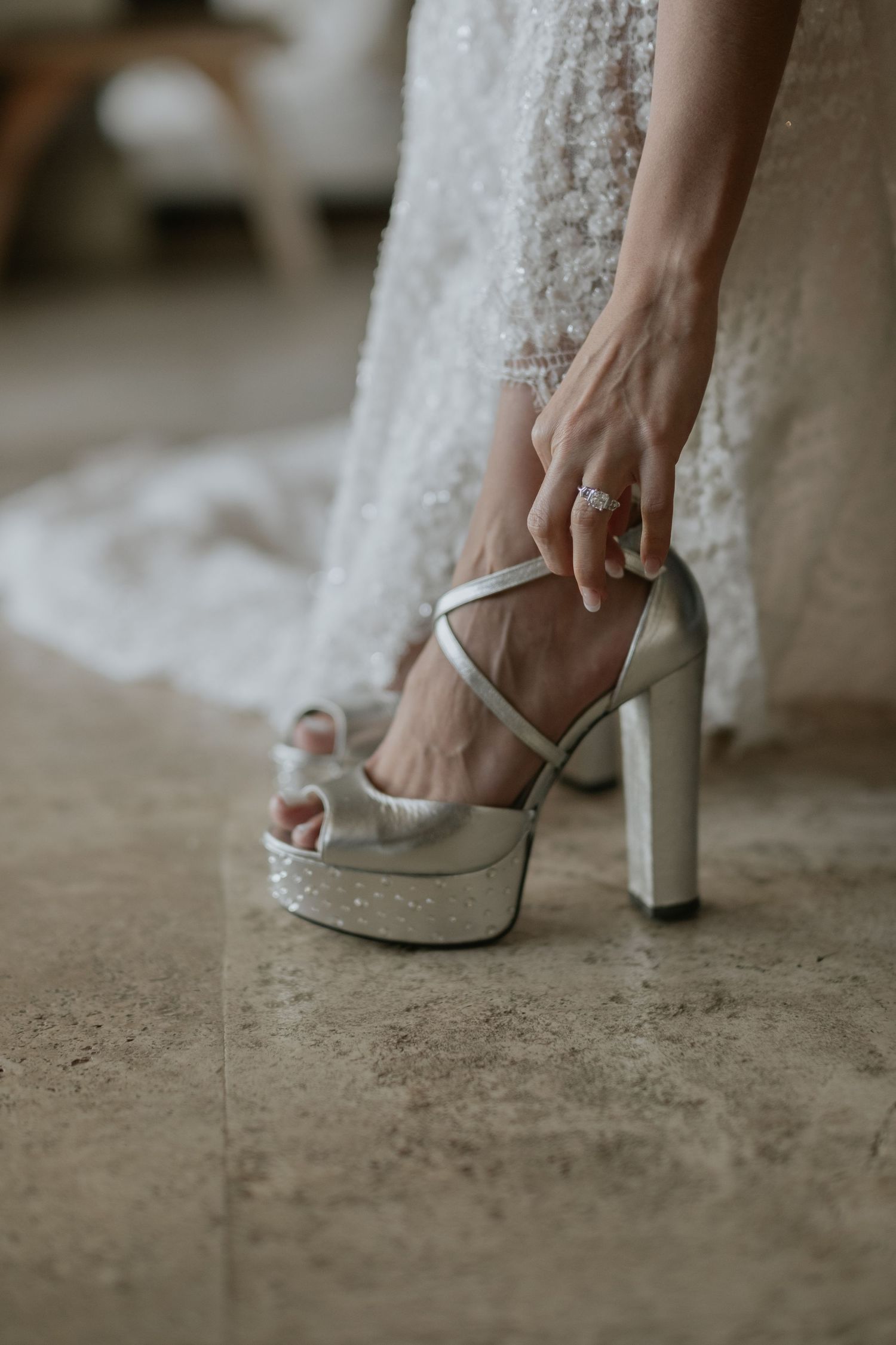 A close-up of a bride's hand fastening elegant silver platform heels with a lace wedding dress in the background.