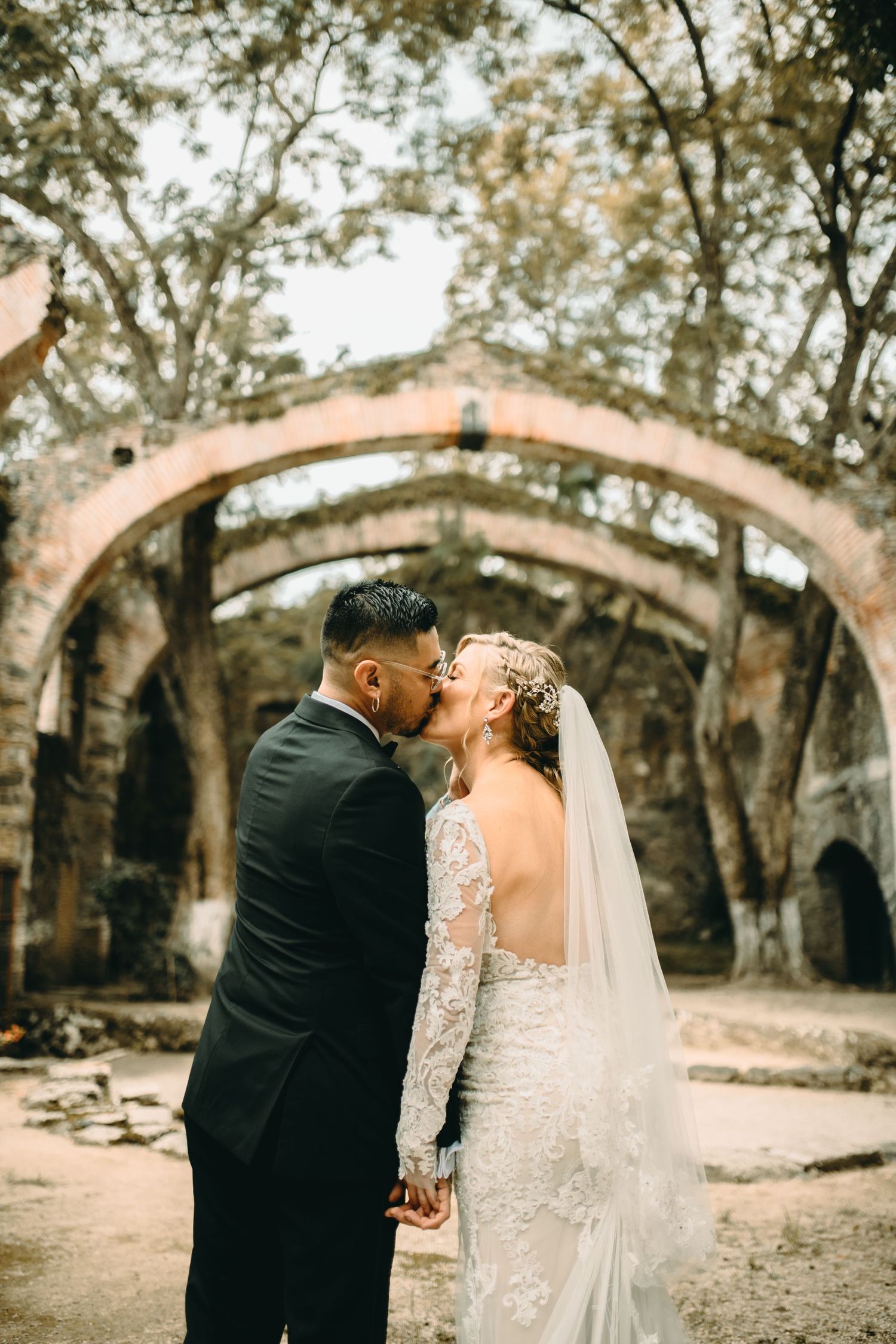 Romantic newlyweds share a kiss under rustic stone arches at an old ruins surrounded by trees.
