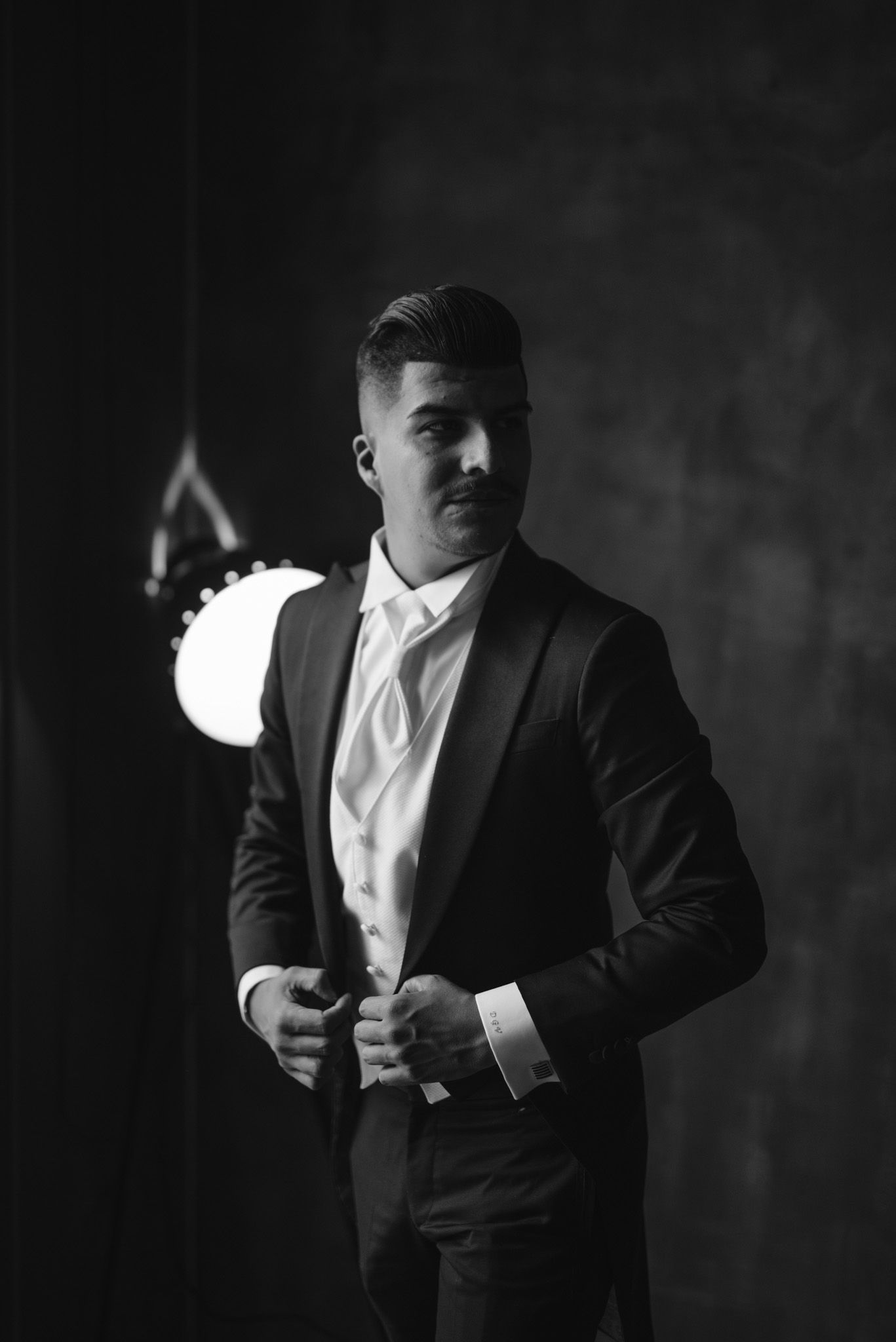 Black and white portrait of person in formal attire adjusting suit jacket in moody lighting.