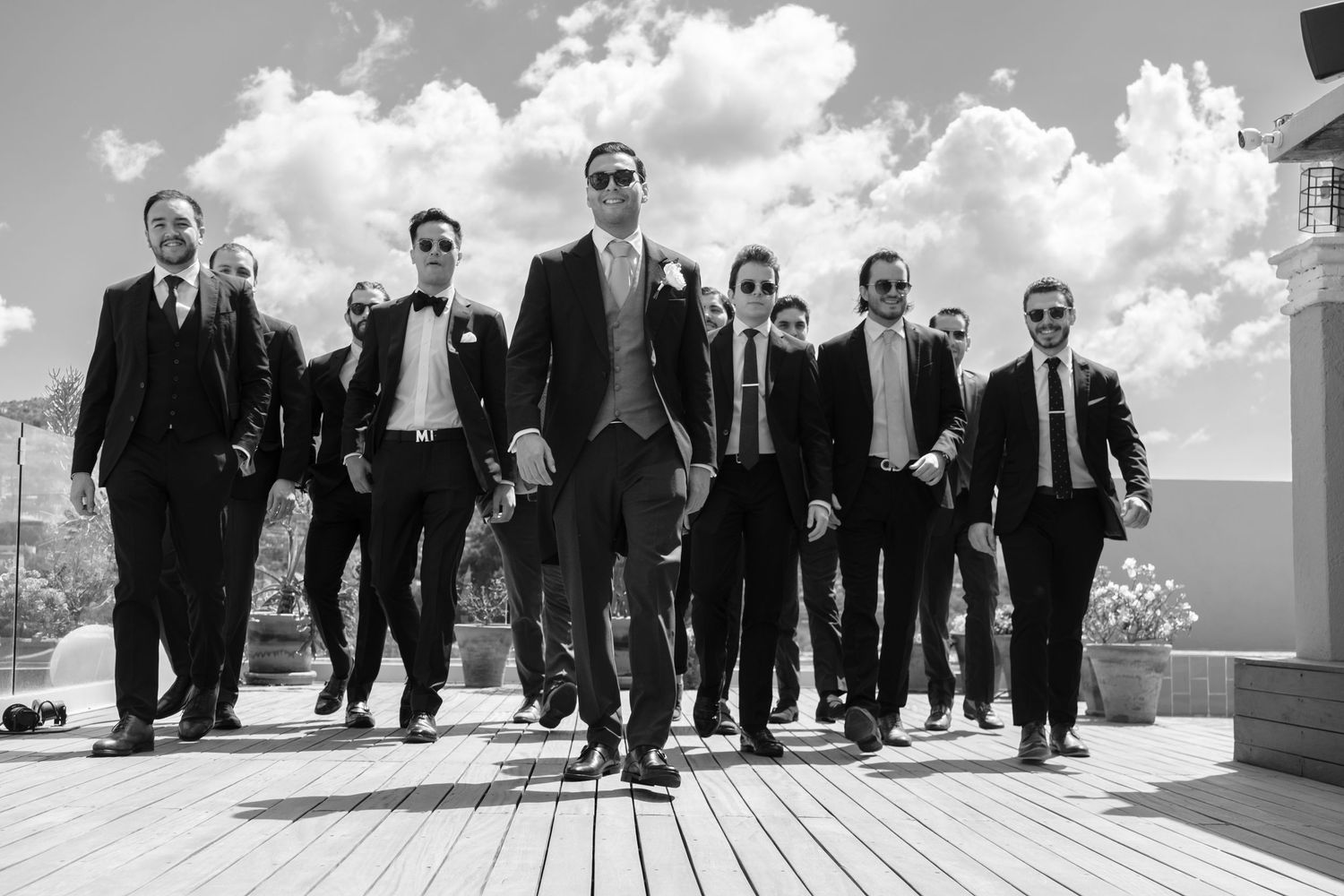 Group of groomsmen walking in black suits against cloudy sky on wooden deck in dramatic black and white photo.