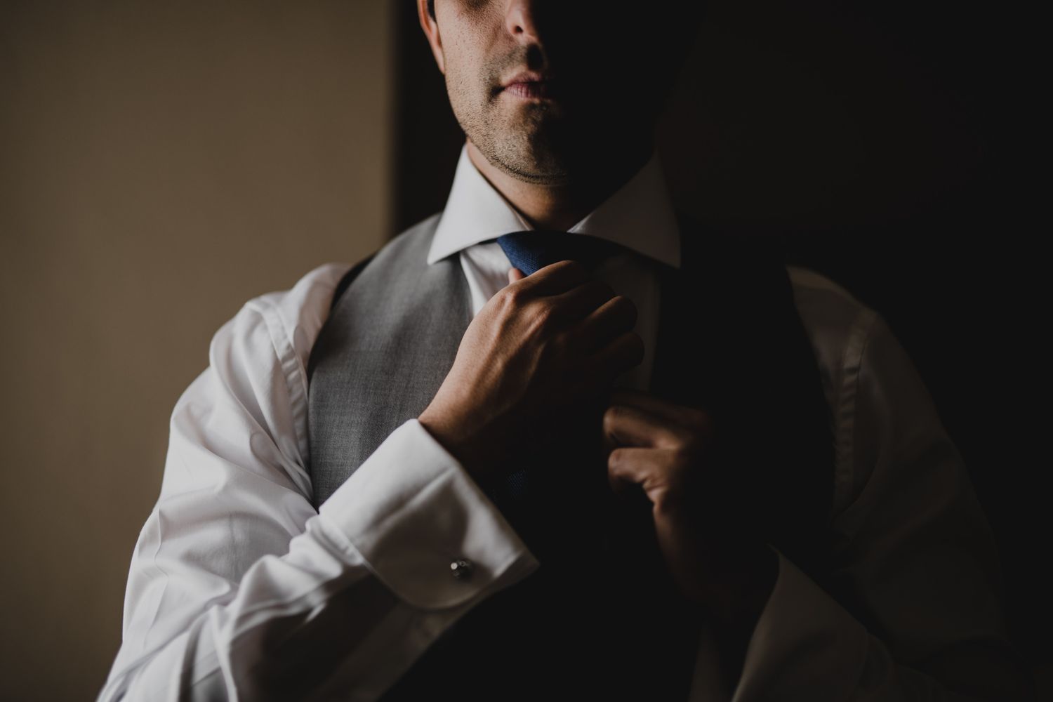 Person adjusting a blue necktie while wearing a gray vest and white dress shirt in moody lighting.
