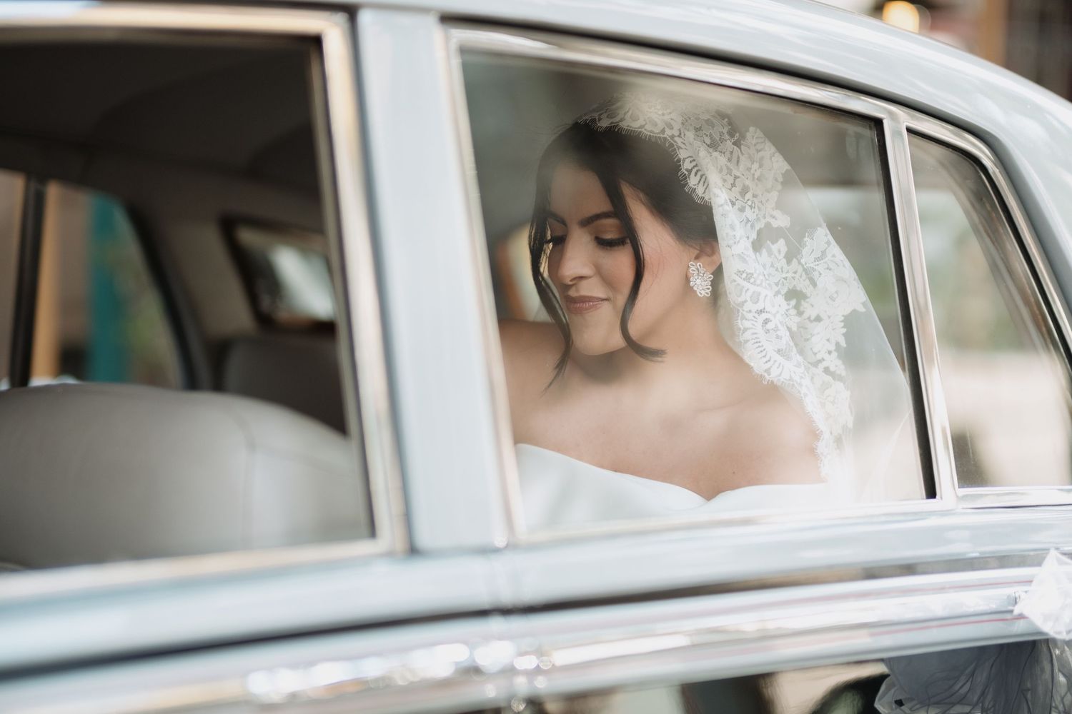 Beautiful bride wearing white veil looks out window of classic car on her wedding day.