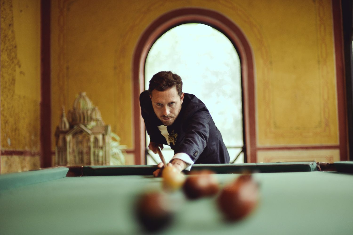 A person in a black jacket leans over a green pool table, focusing on making a shot in a room with yellow walls.