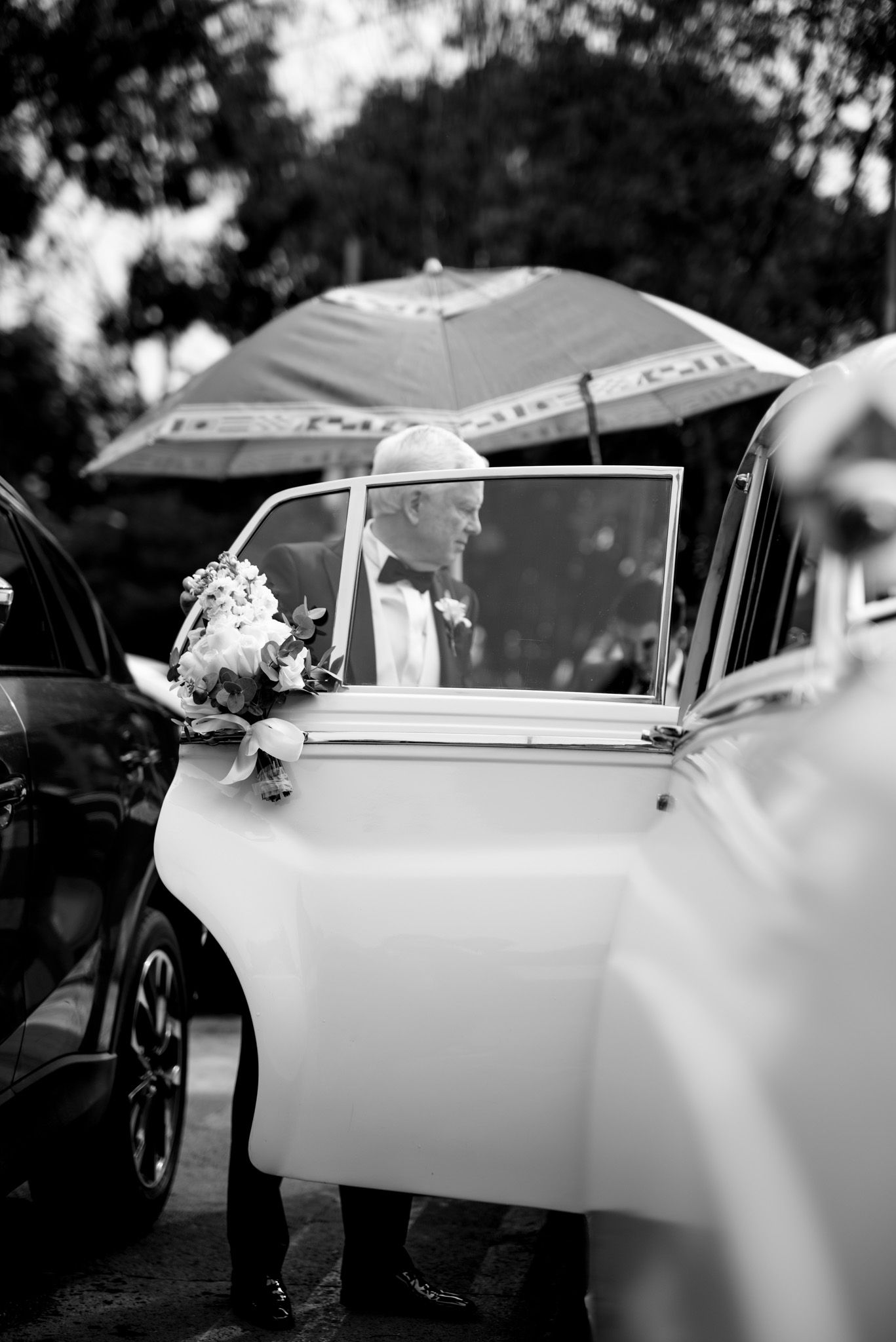 Black and white photo of someone getting into a vintage car with an umbrella and bouquet on a rainy wedding day.