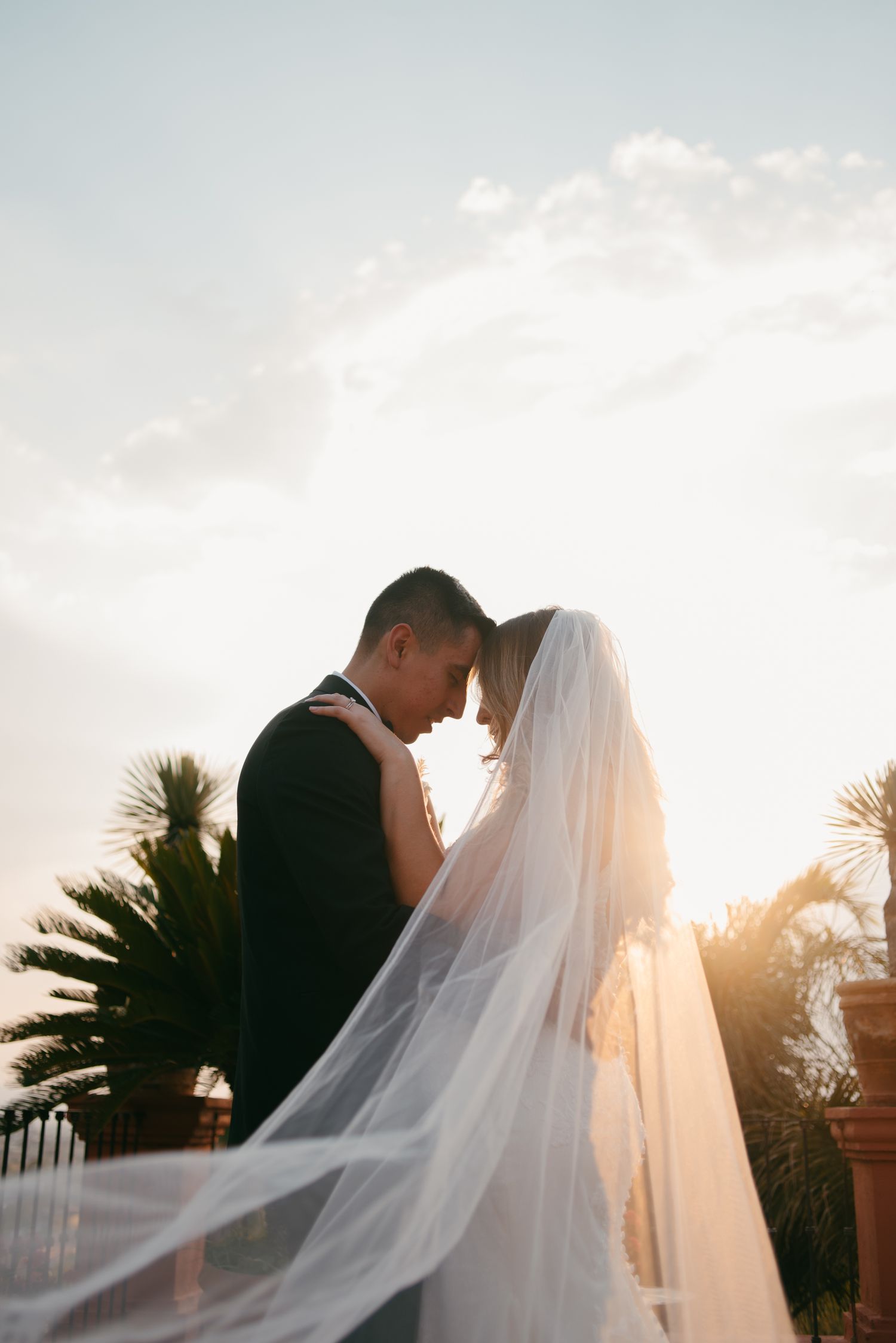 Backlit newlyweds embracing against sunset sky with flowing veil.
