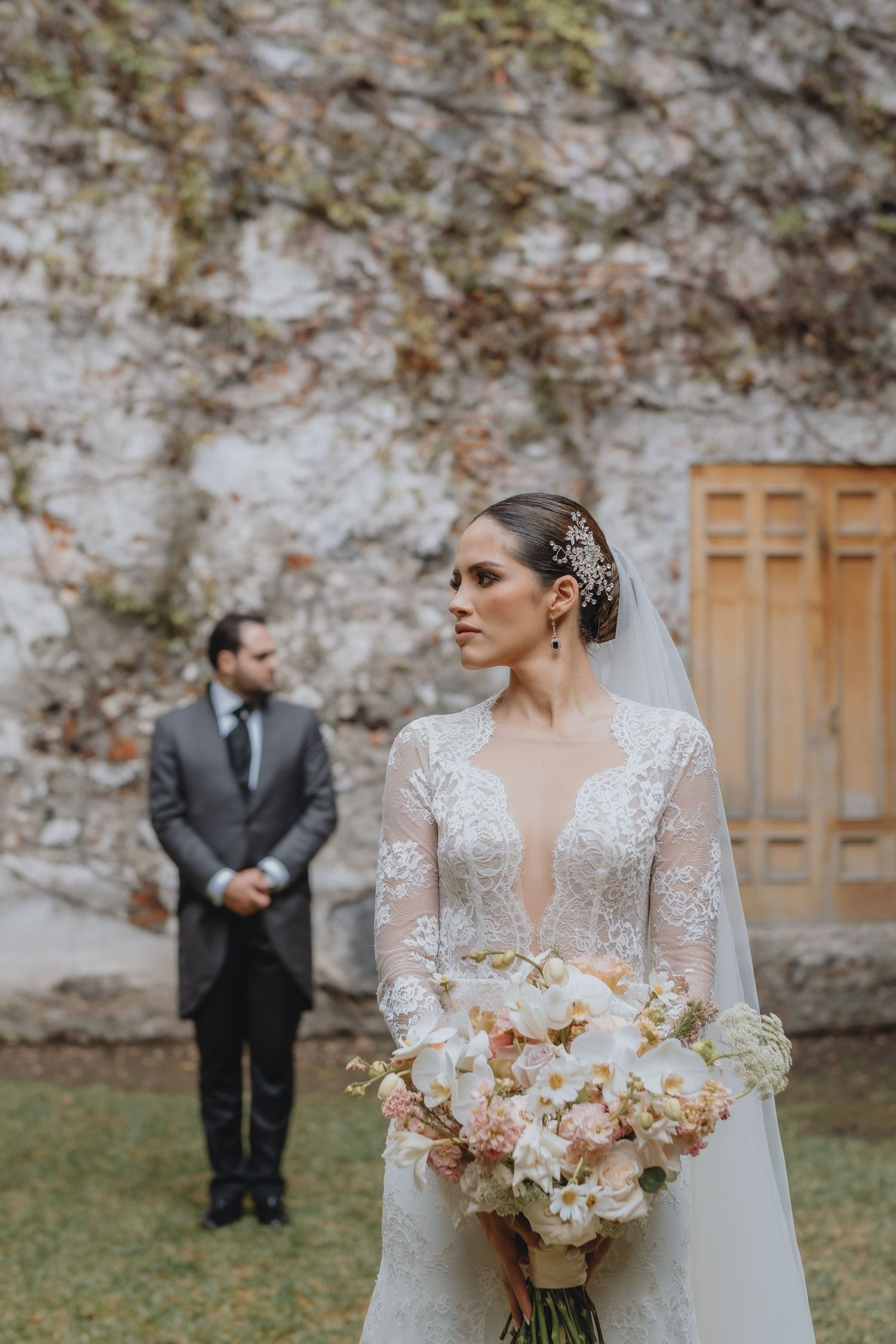 Bride holding peach bouquet in foreground with groom standing in background by stone wall.