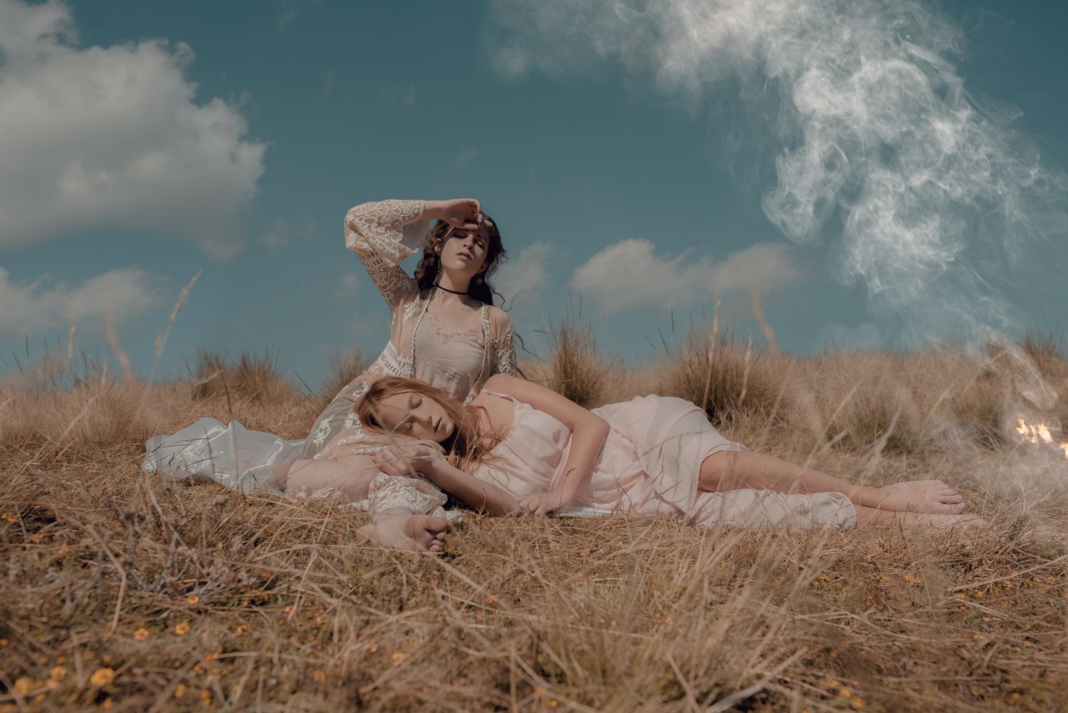 A person in a light dress lies in a golden field under a blue sky with clouds.