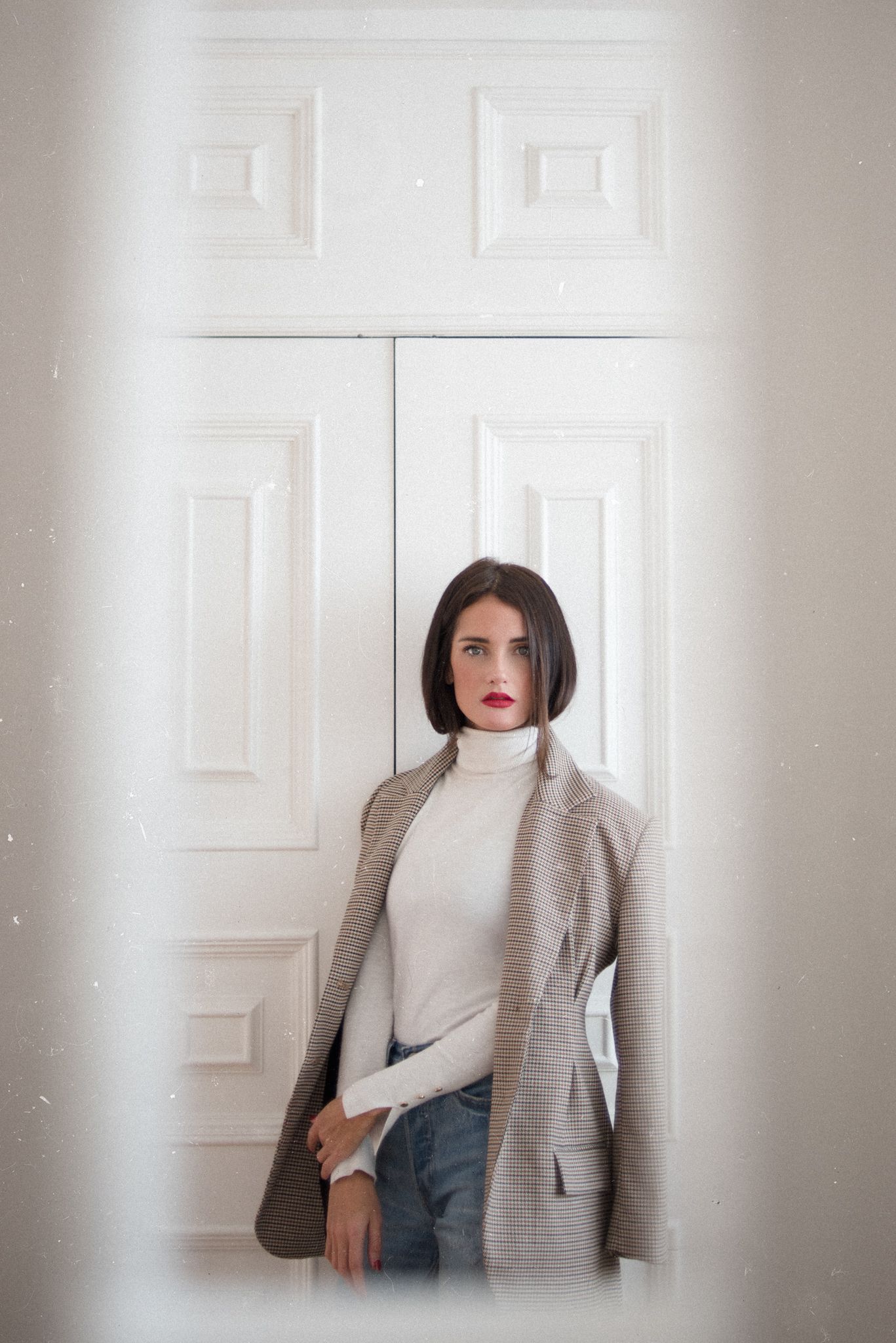 Stylish minimal portrait against white door wearing turtleneck and plaid blazer.