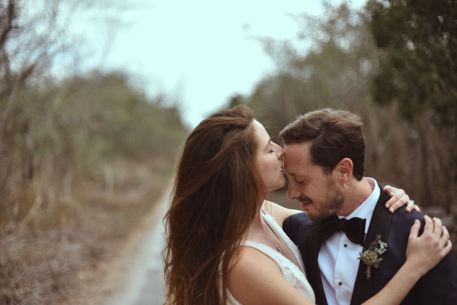 A romantic couple shares an intimate moment together on a scenic outdoor path in formal wedding attire.