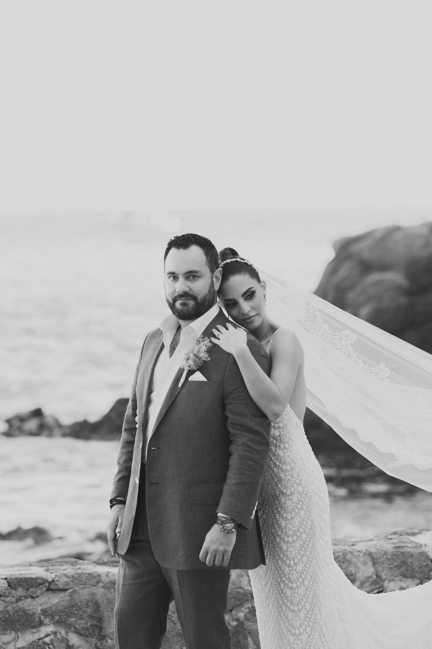 Black and white photo of a couple posing together on rocky coastal cliffs as a wedding veil flows in the breeze.