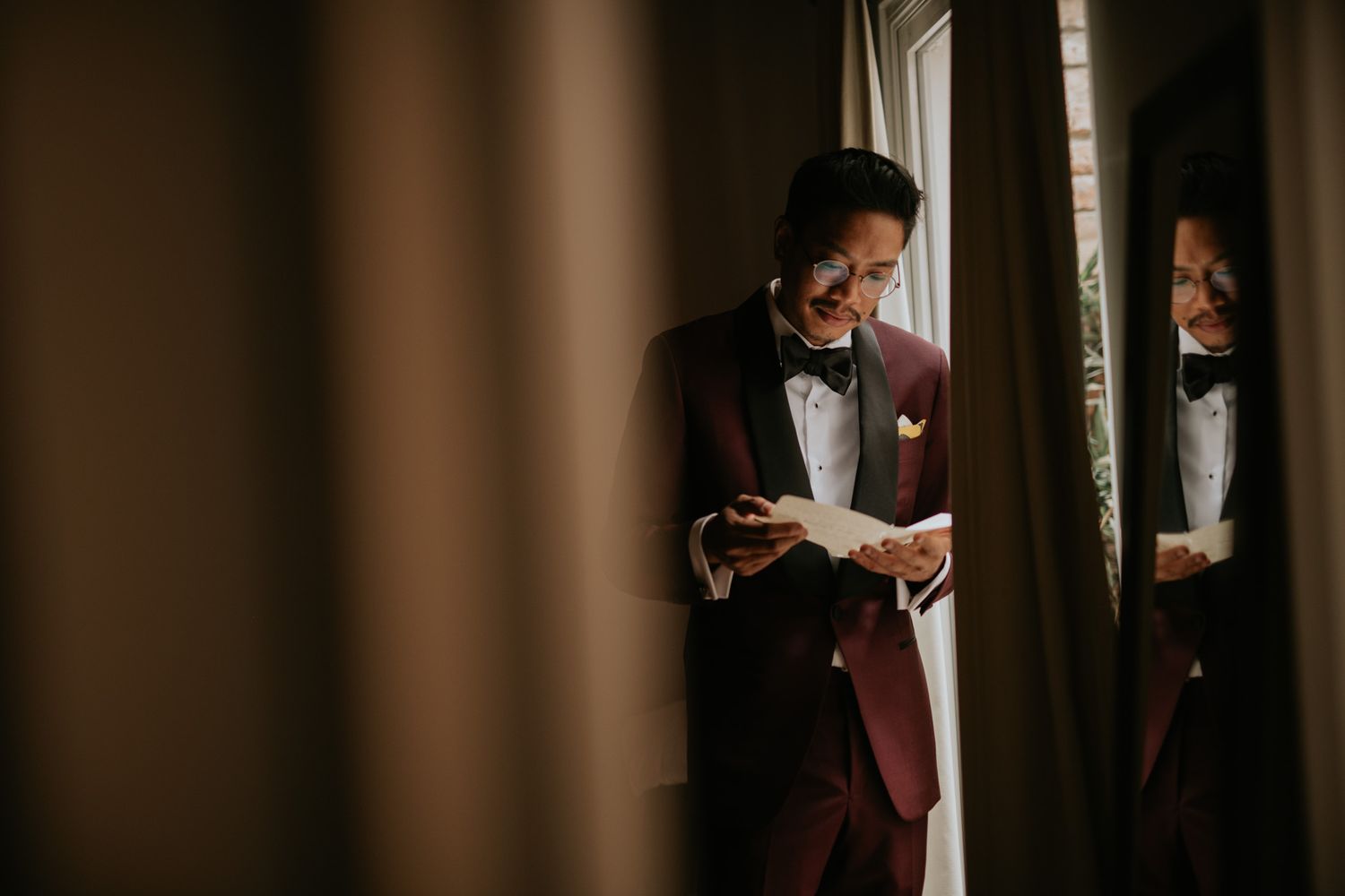 A person in a formal tuxedo looks down at a letter while standing near a window in warm dramatic lighting.