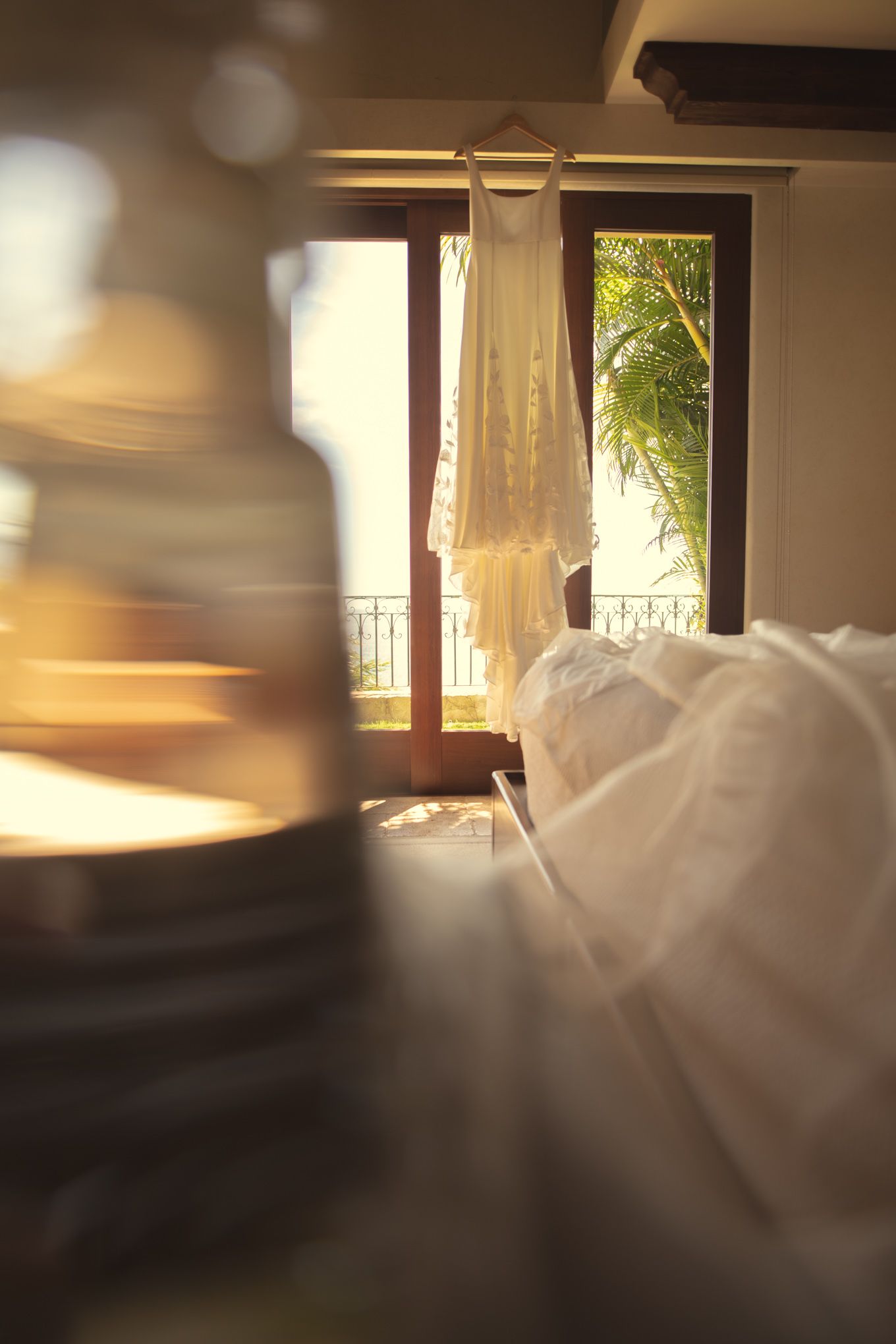 Wedding dress hanging in sunlit room with white bedding and tropical view through balcony window.
