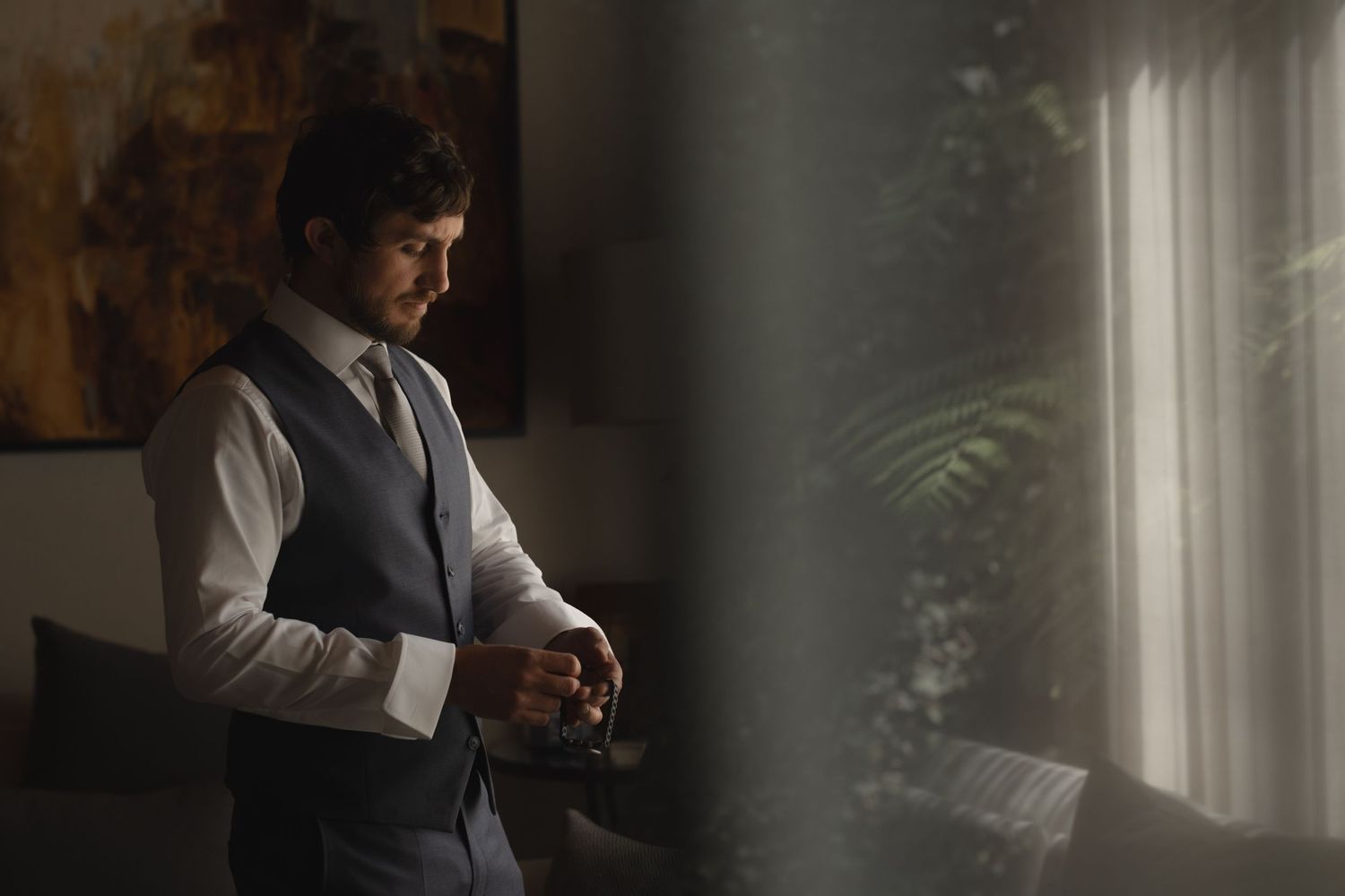 A groom in formal attire checks his watch while standing near a sunlit window.