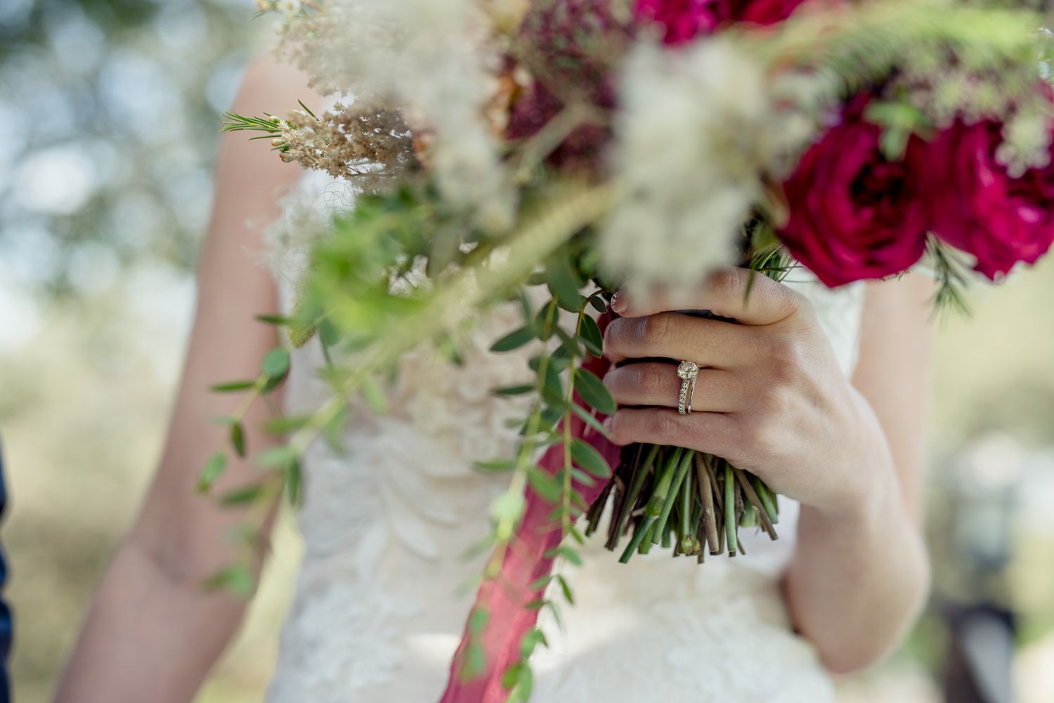 Close-up of bride's hands holding wedding bouquet with red roses and delicate greenery.