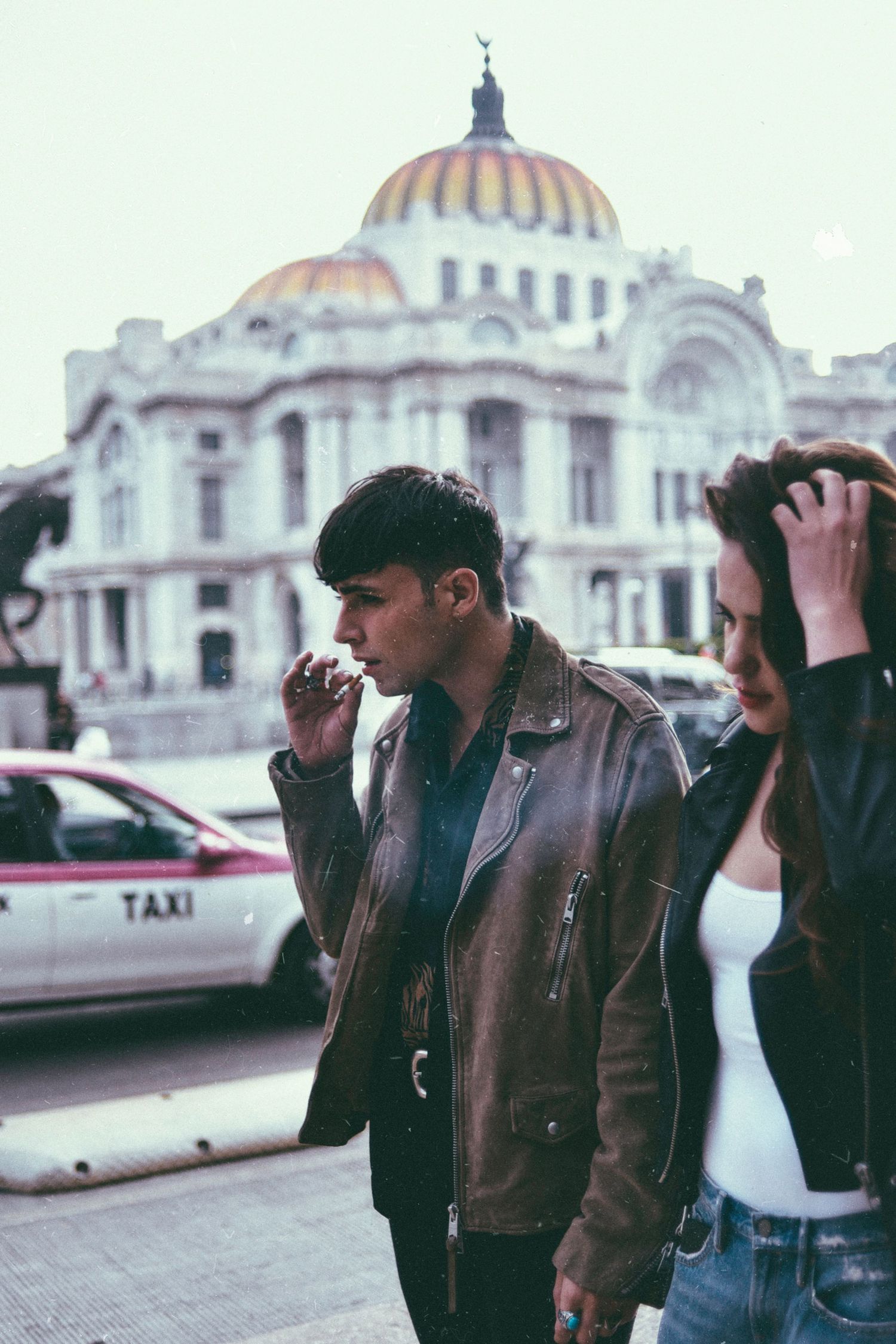 People in leather jackets stand outside Palacio de Bellas Artes with taxi in background.