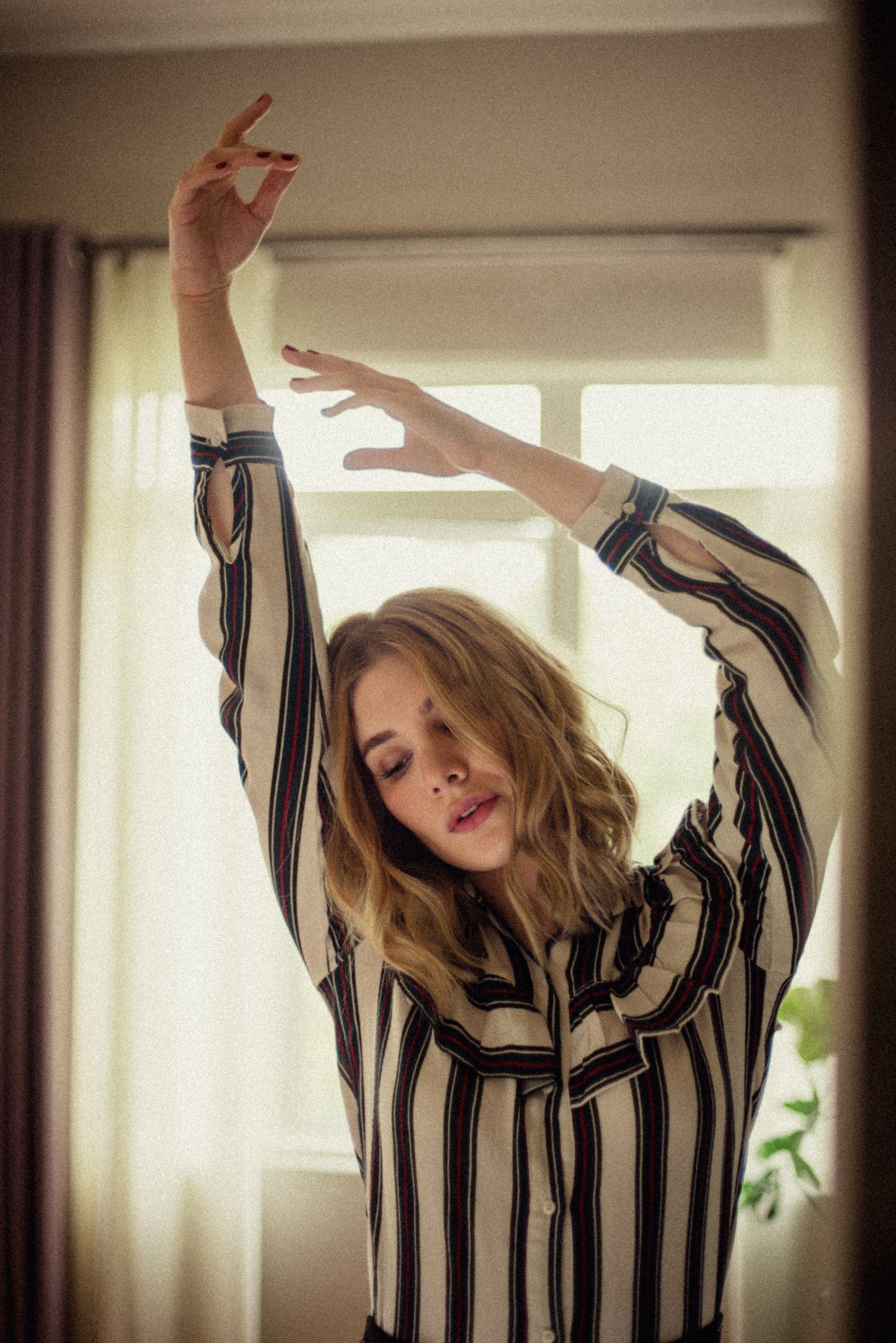 Dancing figure in striped shirt captured in soft window light with raised arms.