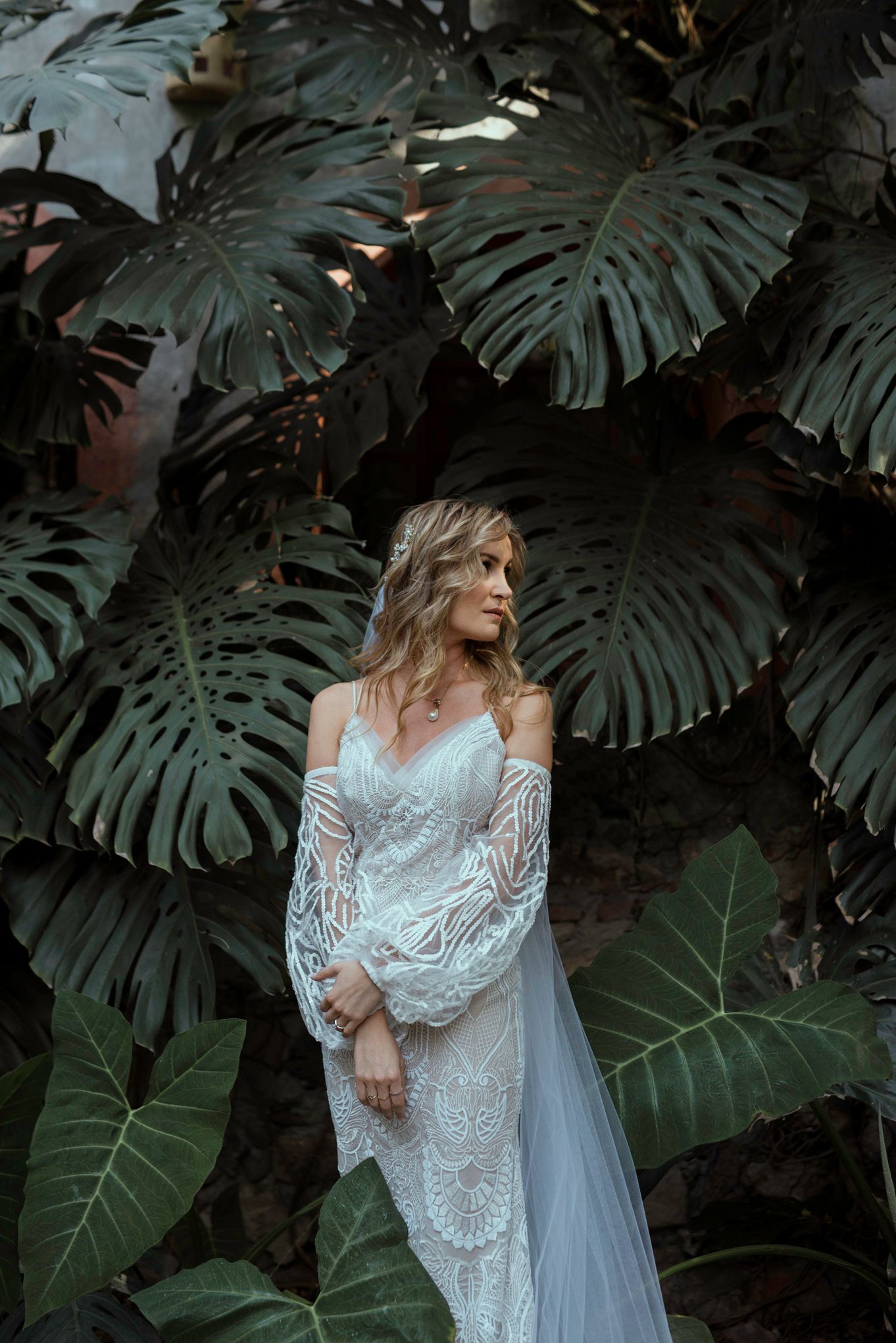 Bride in lace gown poses against tropical monstera leaves in moody lighting.