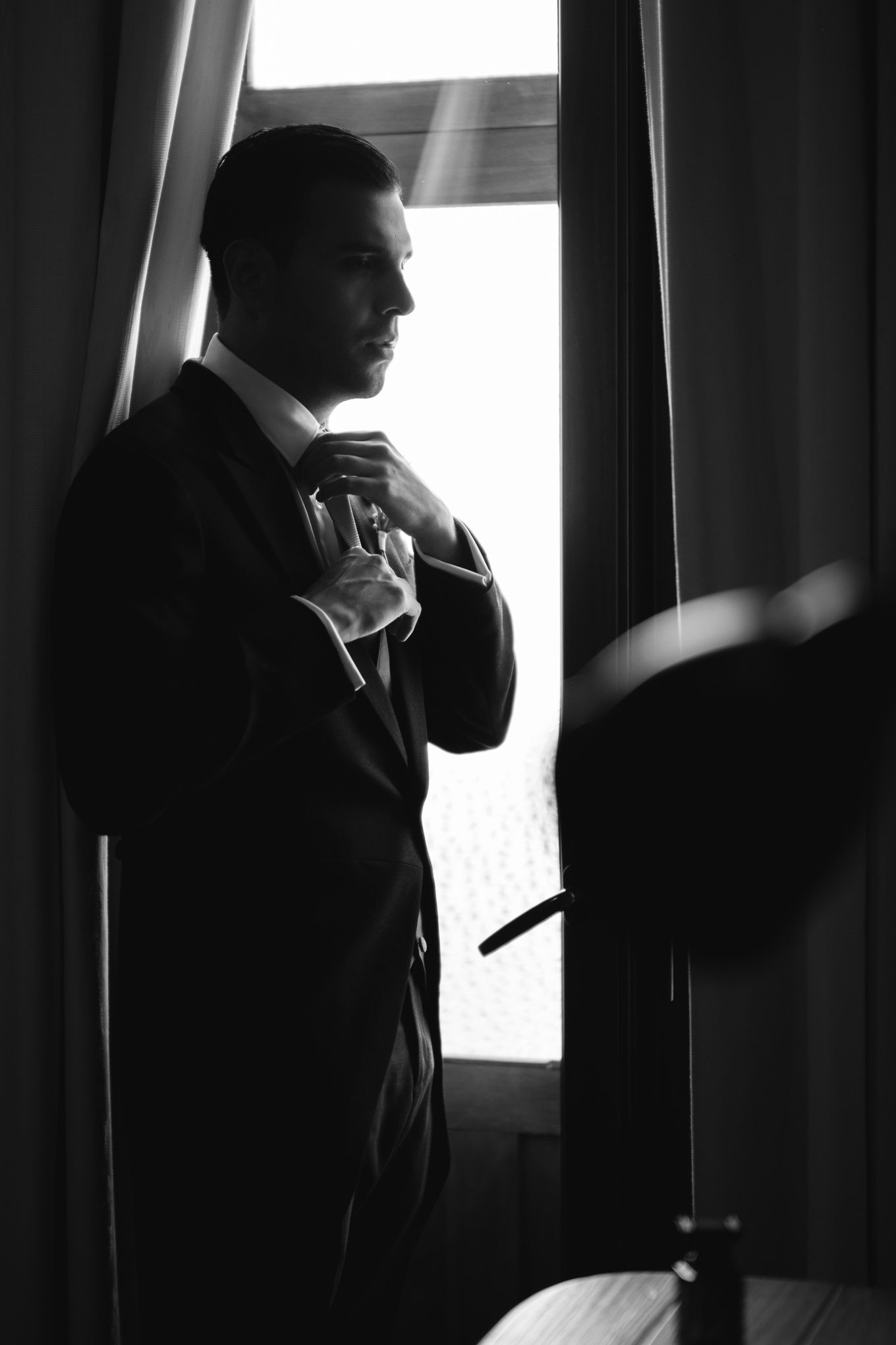 Silhouette of groom adjusting tie while standing by window in dramatic lighting.