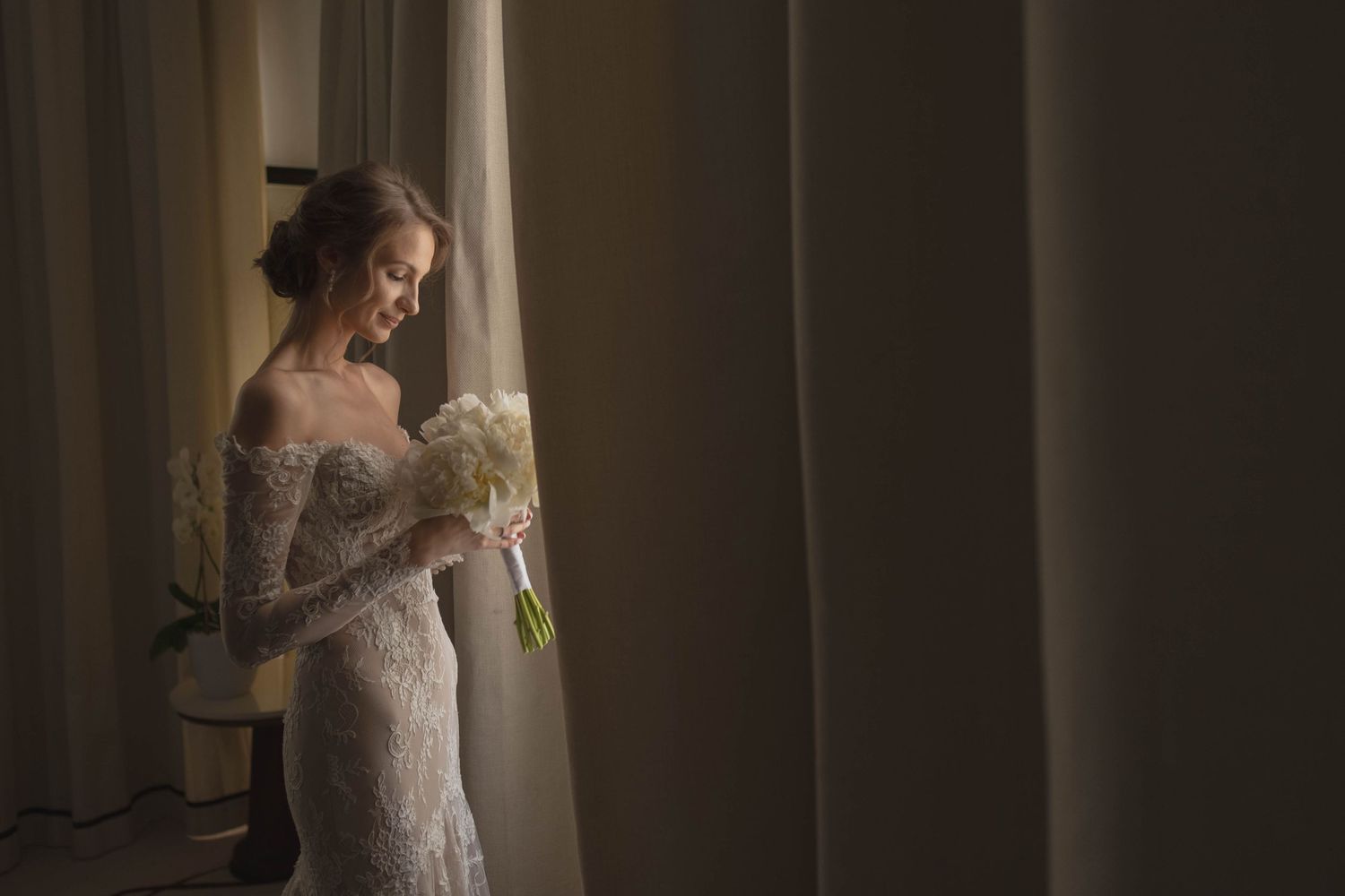 A bride in an elegant lace gown poses by a window in dramatic lighting.