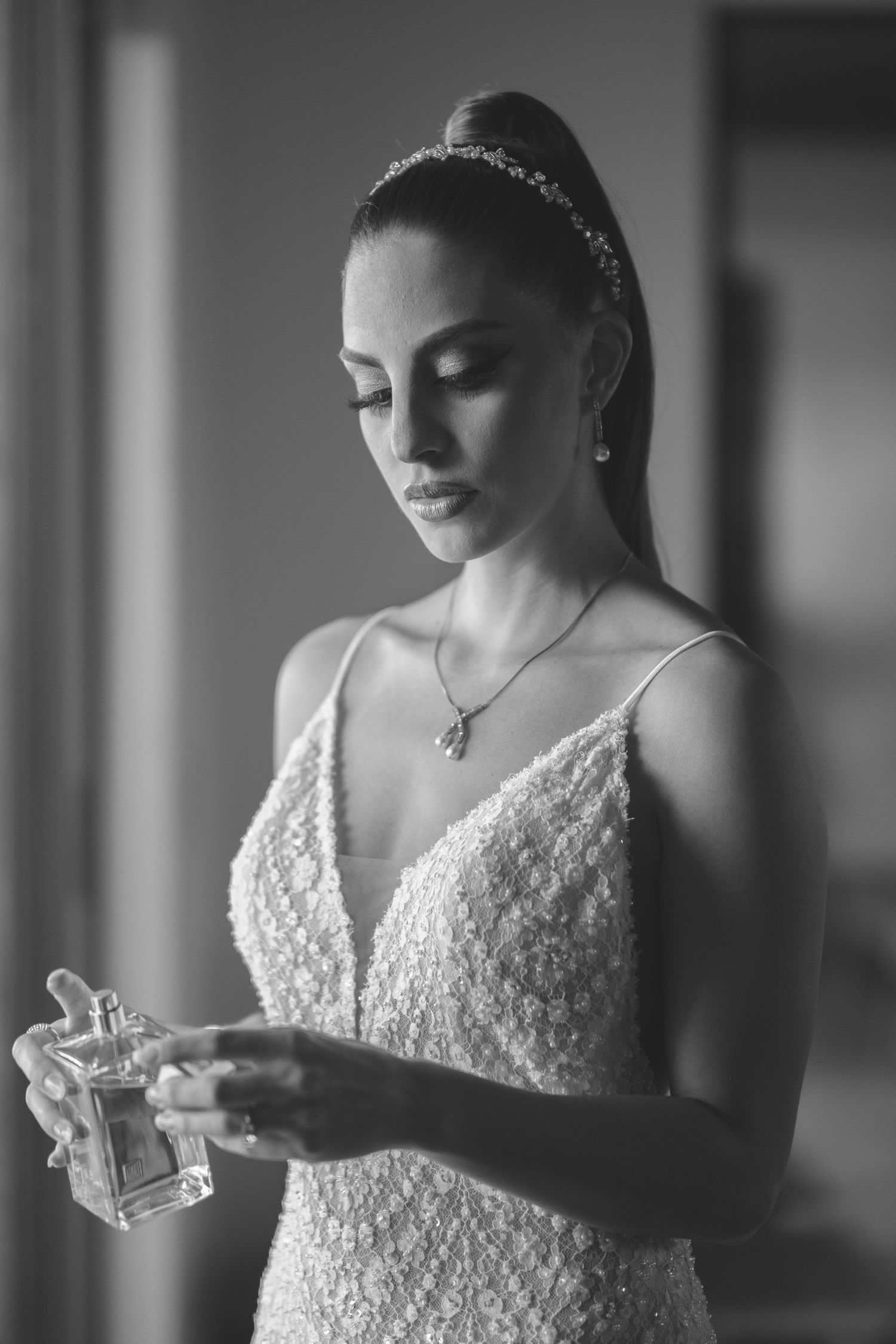 A bride in a lace wedding dress holds a perfume bottle while getting ready in black and white photography.