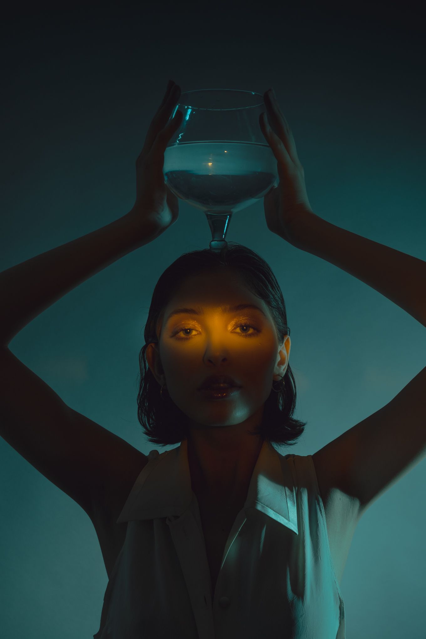 Artistic portrait of model holding a wine glass overhead in dramatic teal lighting creating a moody atmosphere.