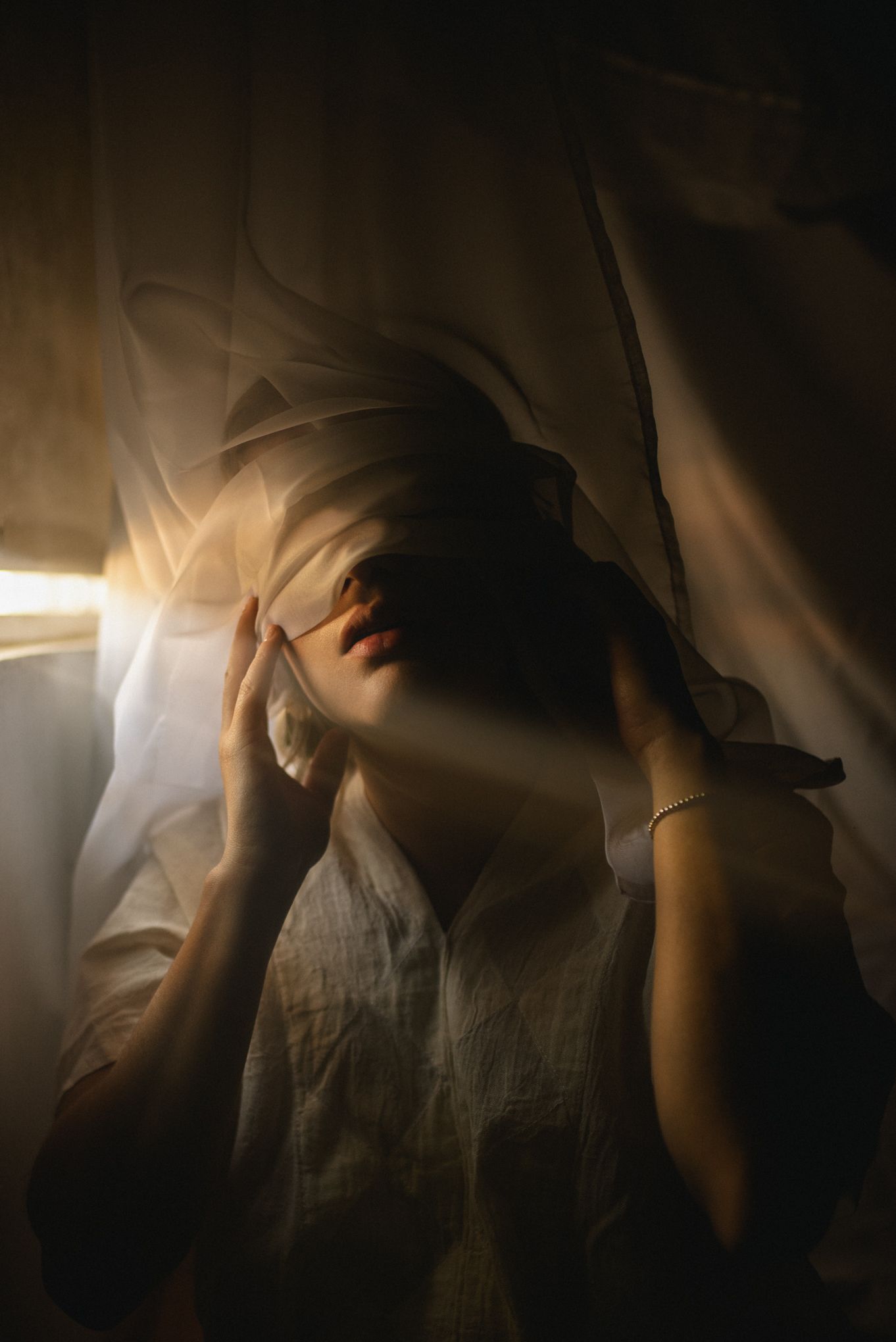 Dramatic low-key portrait with light rays through fabric creating ethereal atmosphere.