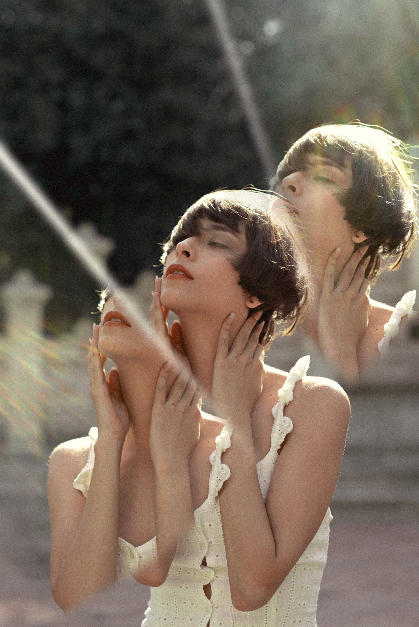 Two women in white dresses pose artistically with hands on their necks in sunlit setting.