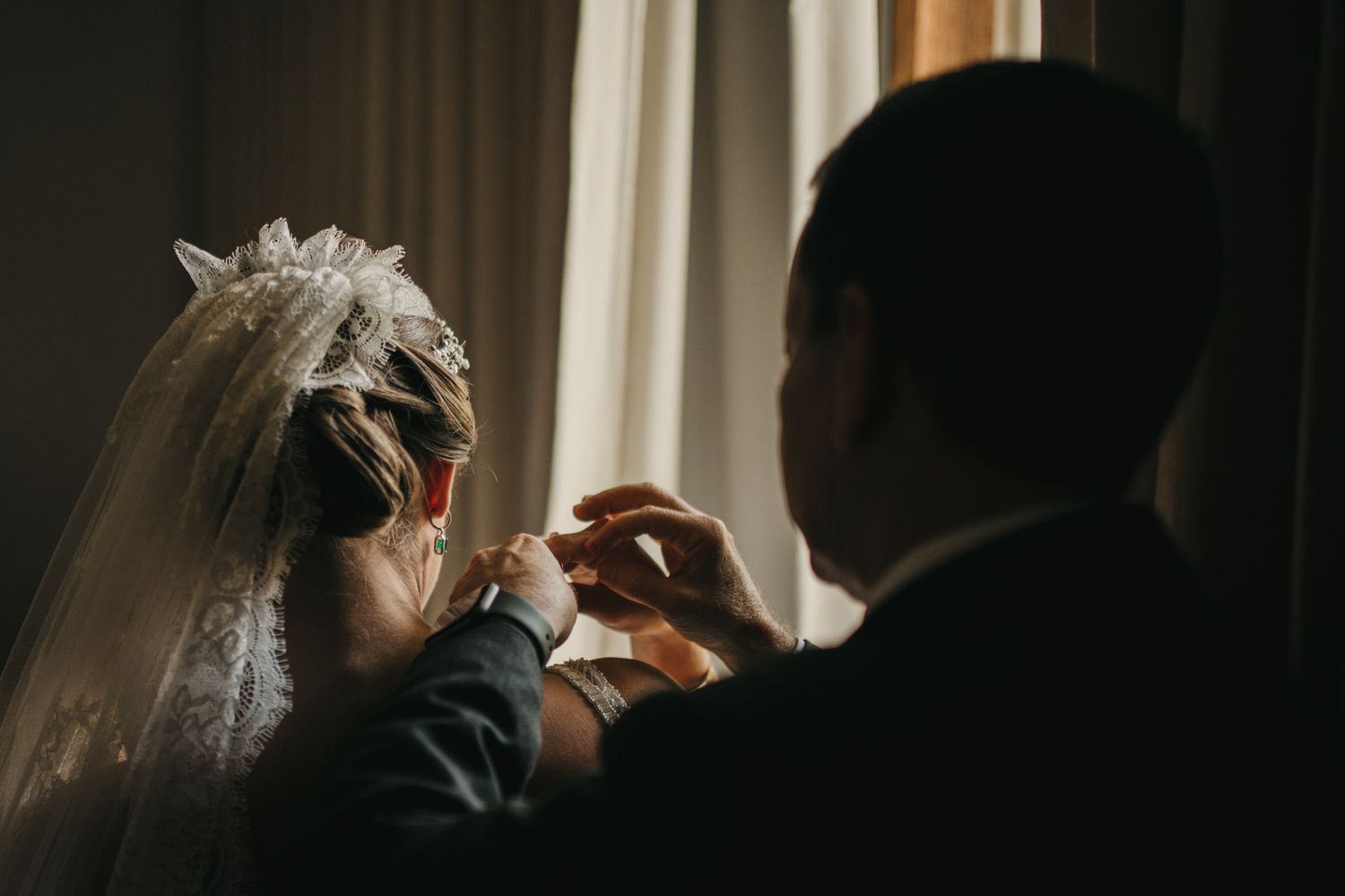 A silhouetted figure gently fixes the veil on a bride in a softly lit room during wedding preparations.