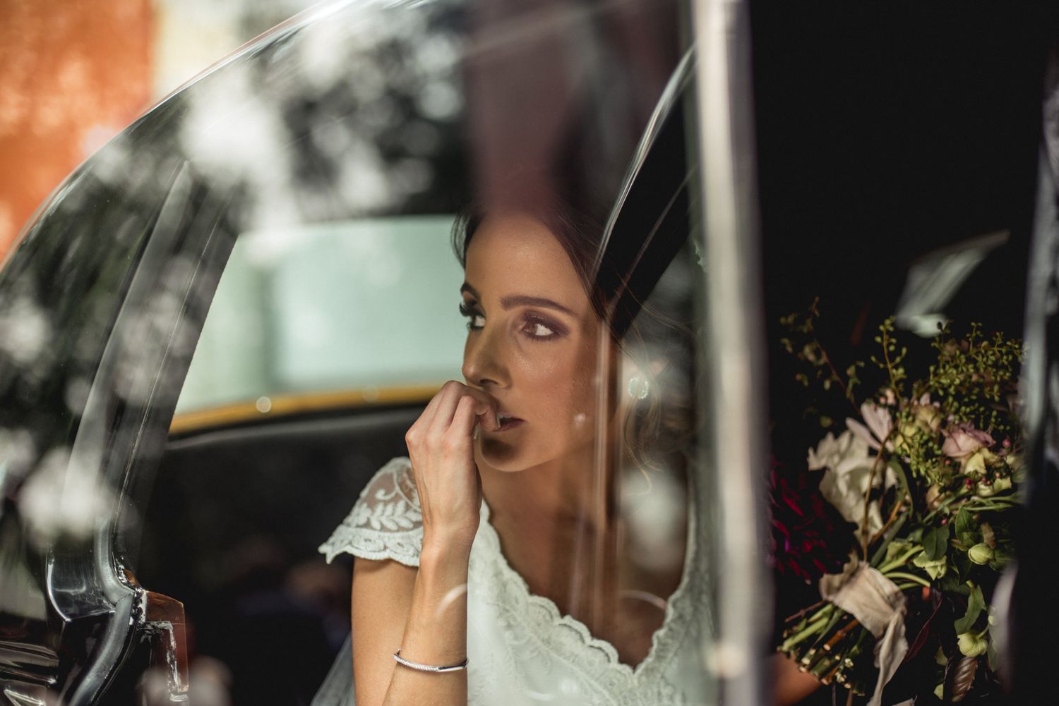 Pensive bride in white lace dress gazes thoughtfully through car window with flowers in background.
